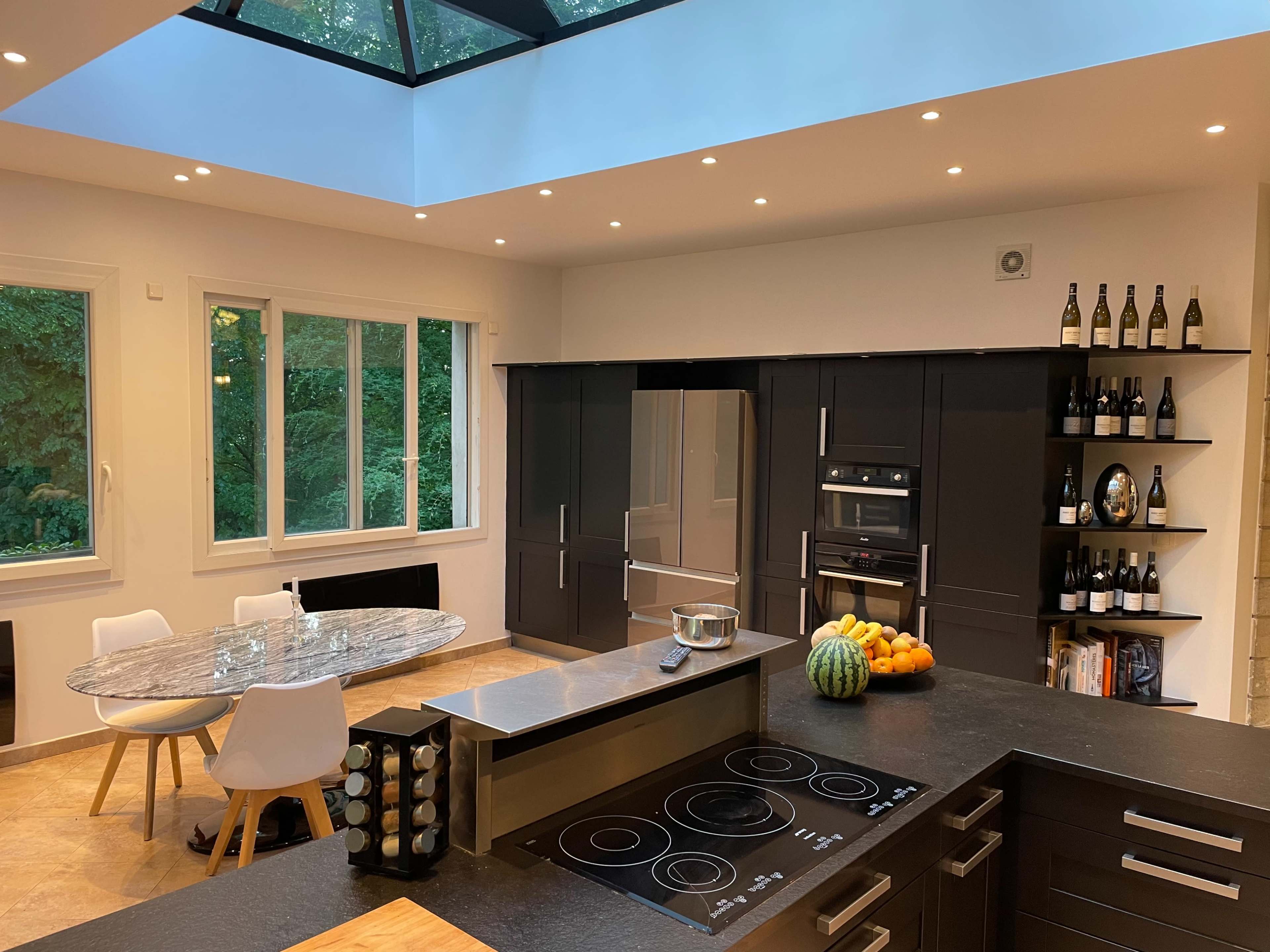The kitchen features a modern design with a central island, a round dining table, and large windows that provide a view of the surrounding greenery.