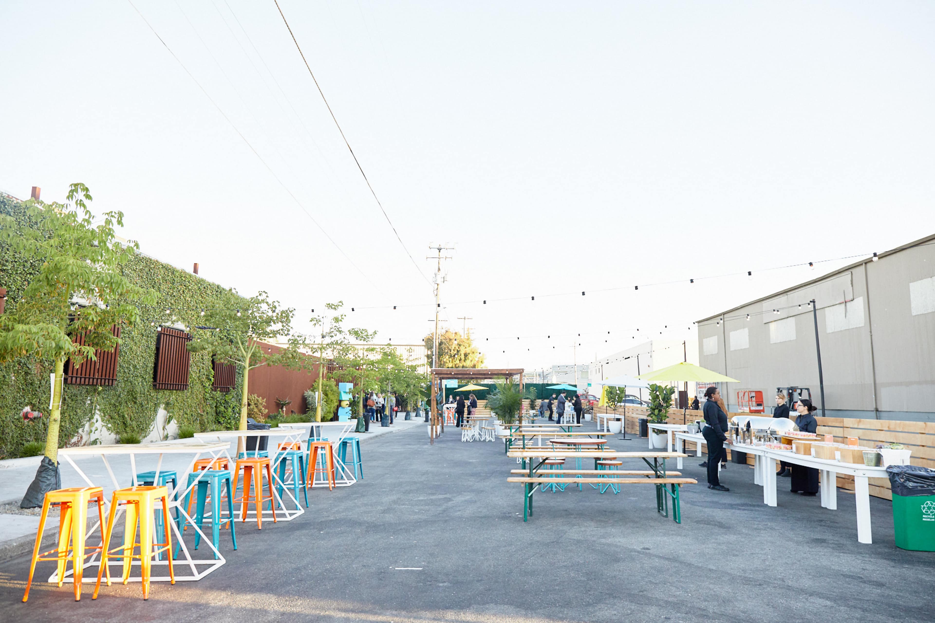 The image shows an outdoor event space with tables, chairs, and people gathering, set against a backdrop of greenery and industrial buildings.