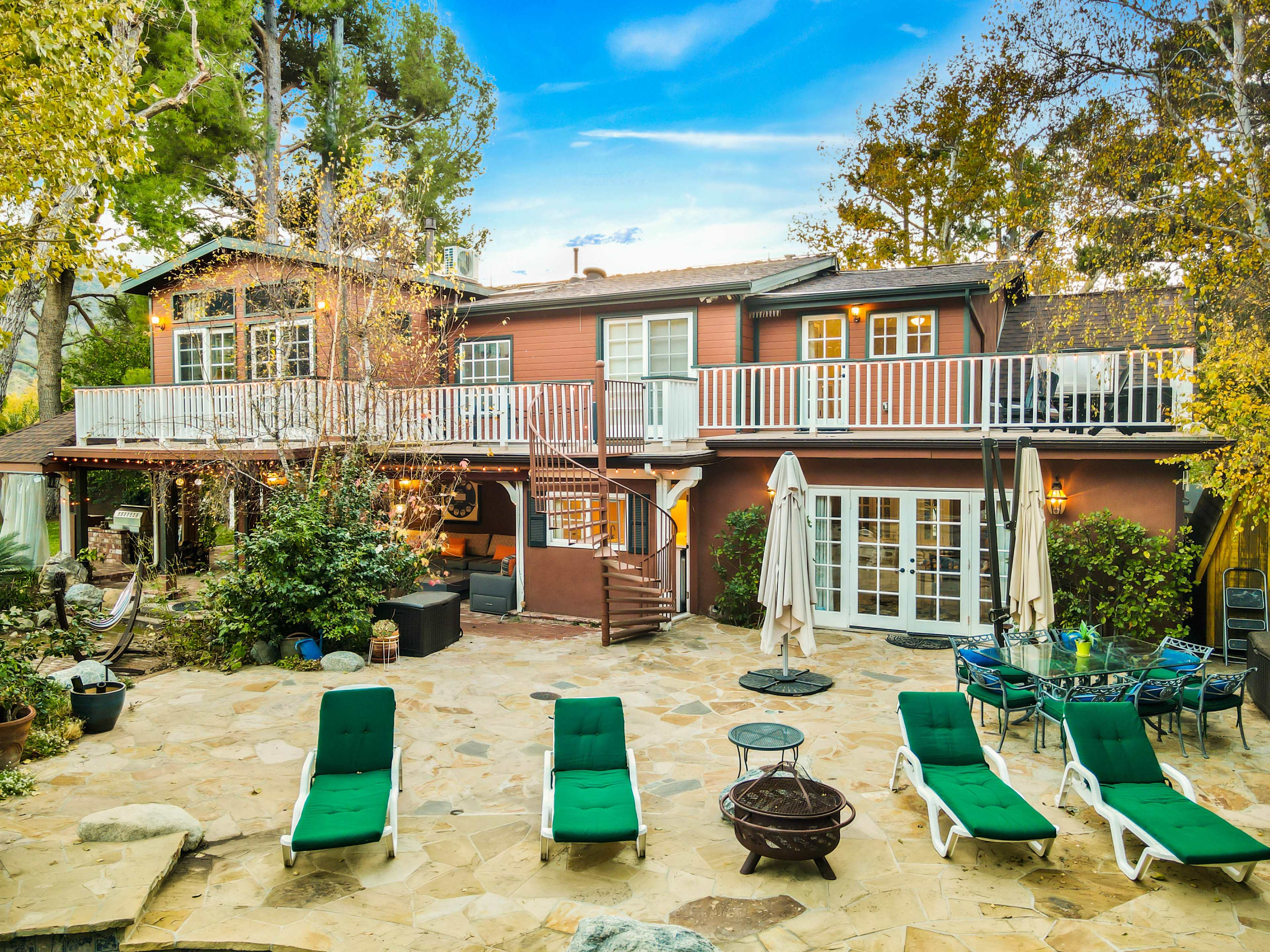 The image shows a multi-level house with a balcony in a landscaped yard featuring lounge chairs and outdoor seating areas.