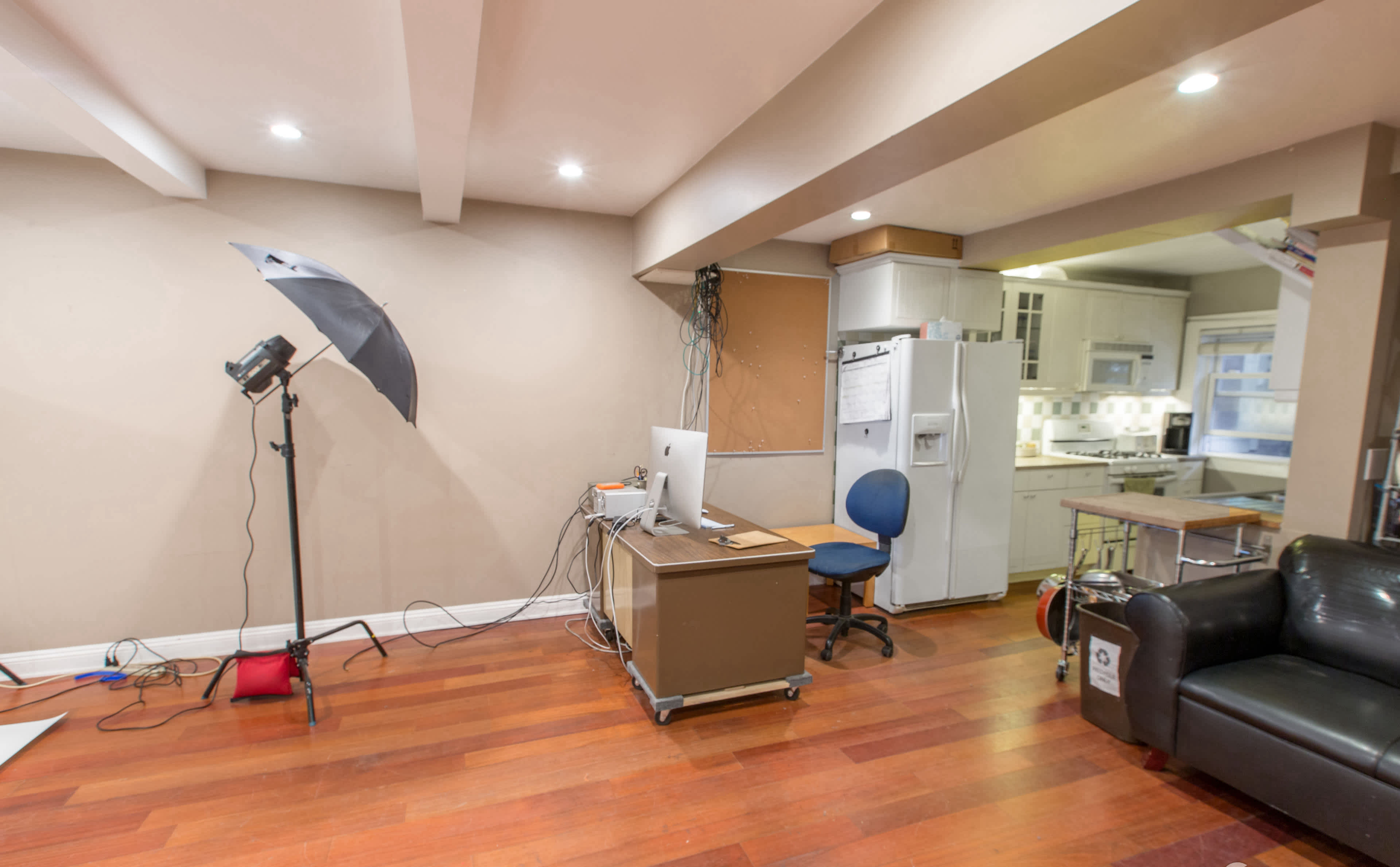 A spacious room with wooden flooring, featuring a desk with a computer, a chair, a softbox light, and a kitchen in the background.