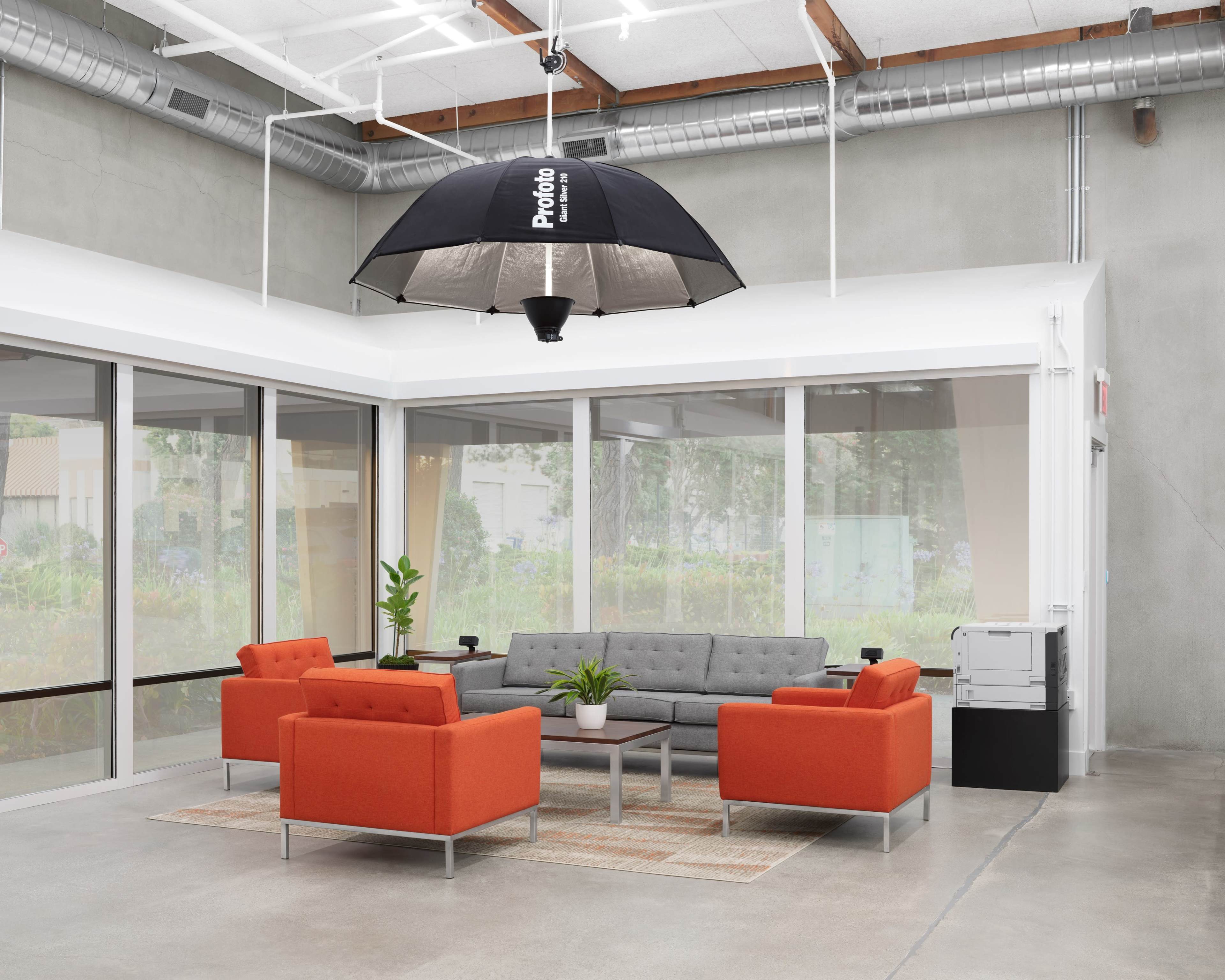 A modern lounge area with a gray sofa, two orange chairs, and a large umbrella light overhead, surrounded by windows.