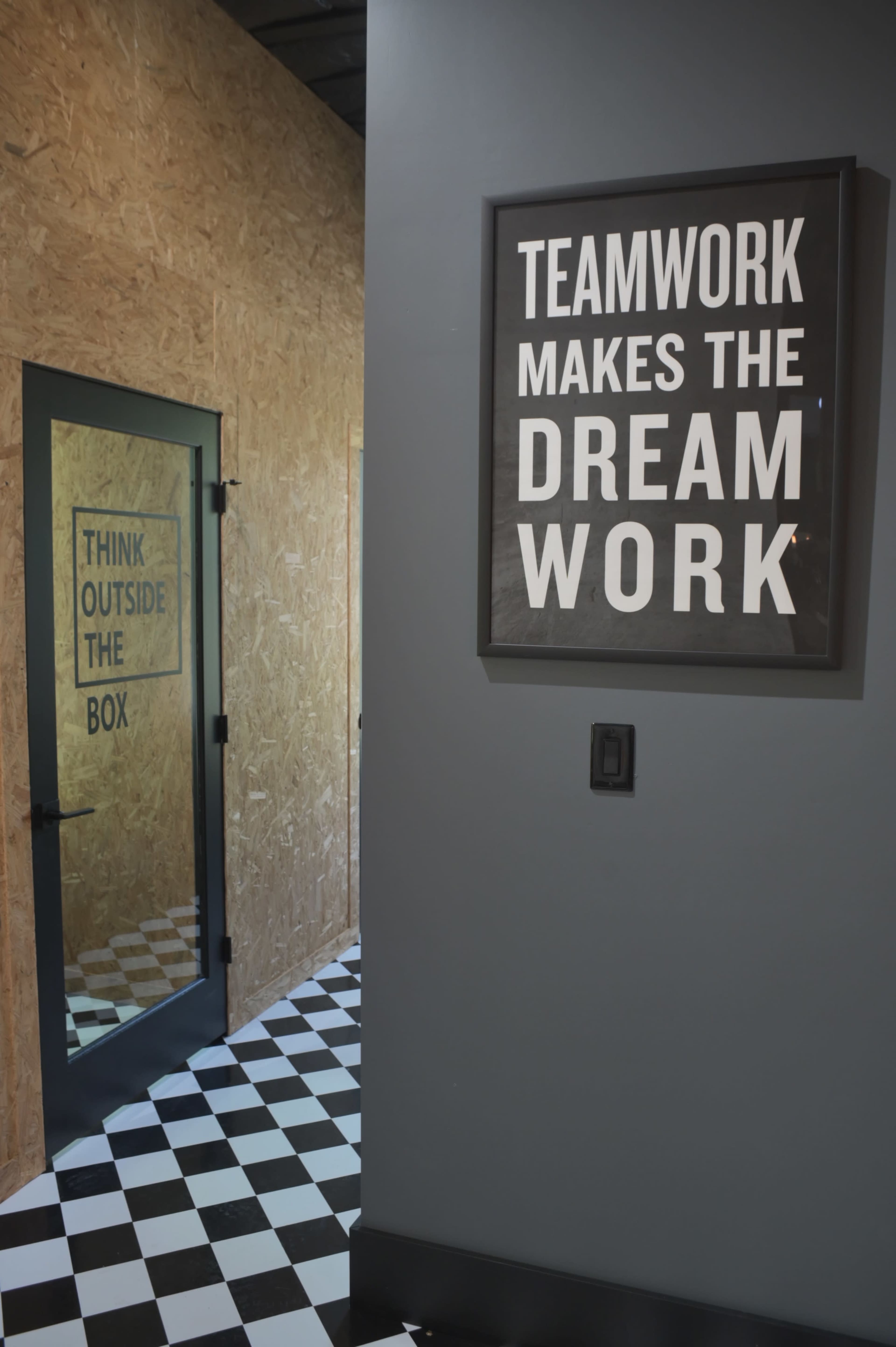 A hallway with a black and white checked floor, featuring a door with "THINK OUTSIDE THE BOX" on the wall opposite a framed sign that reads "TEAMWORK MAKES THE DREAM WORK."