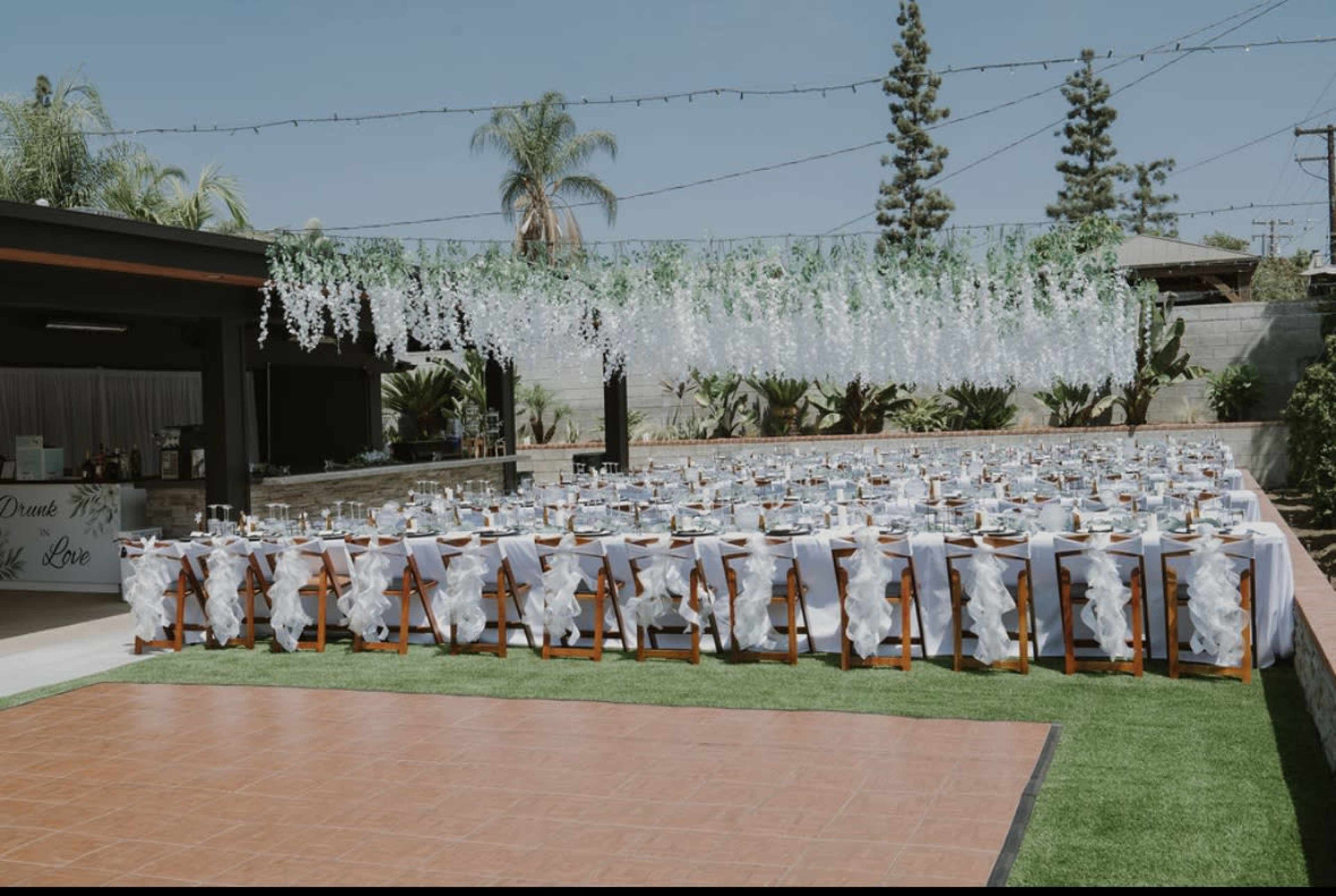 A large outdoor event space features multiple tables arranged for a formal dining setup, adorned with white tablecloths and decorations under a clear blue sky.