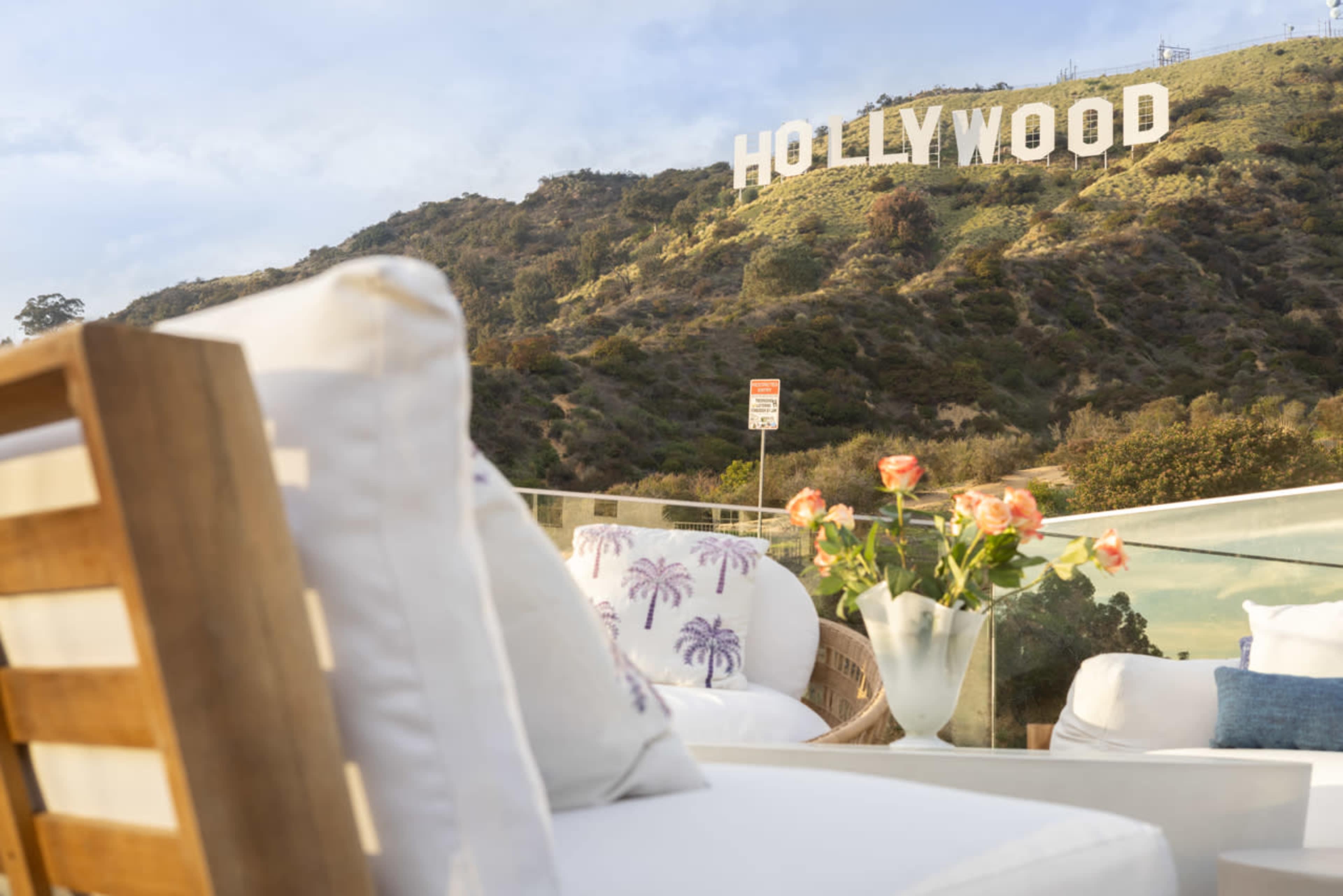 A view from a balcony featuring a comfortable seating area and a vase of flowers, with the Hollywood Sign visible on a hillside in the background.