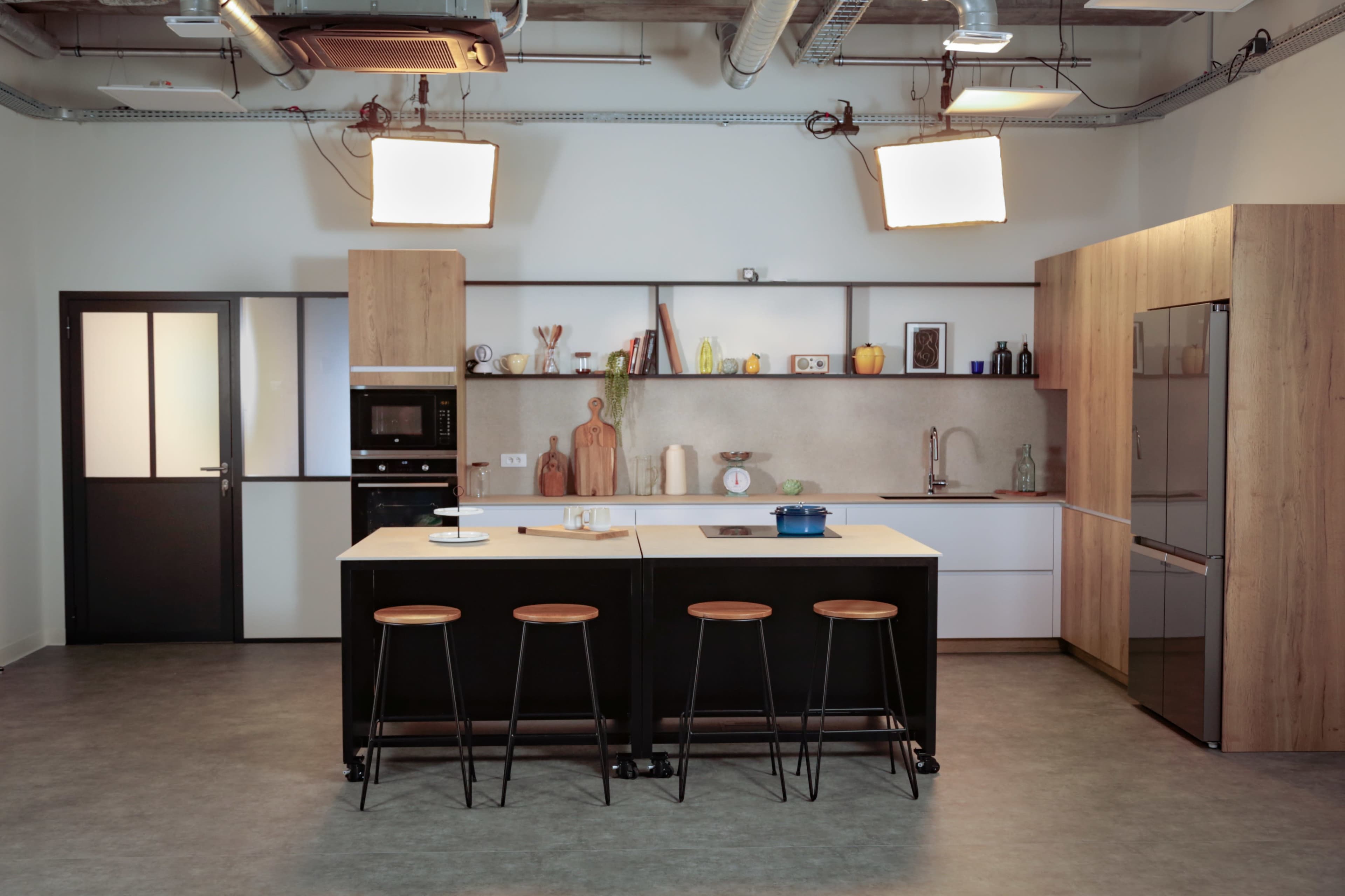 The image shows a modern kitchen with a central island, wooden cabinetry, and various cooking appliances, illuminated by overhead lights.