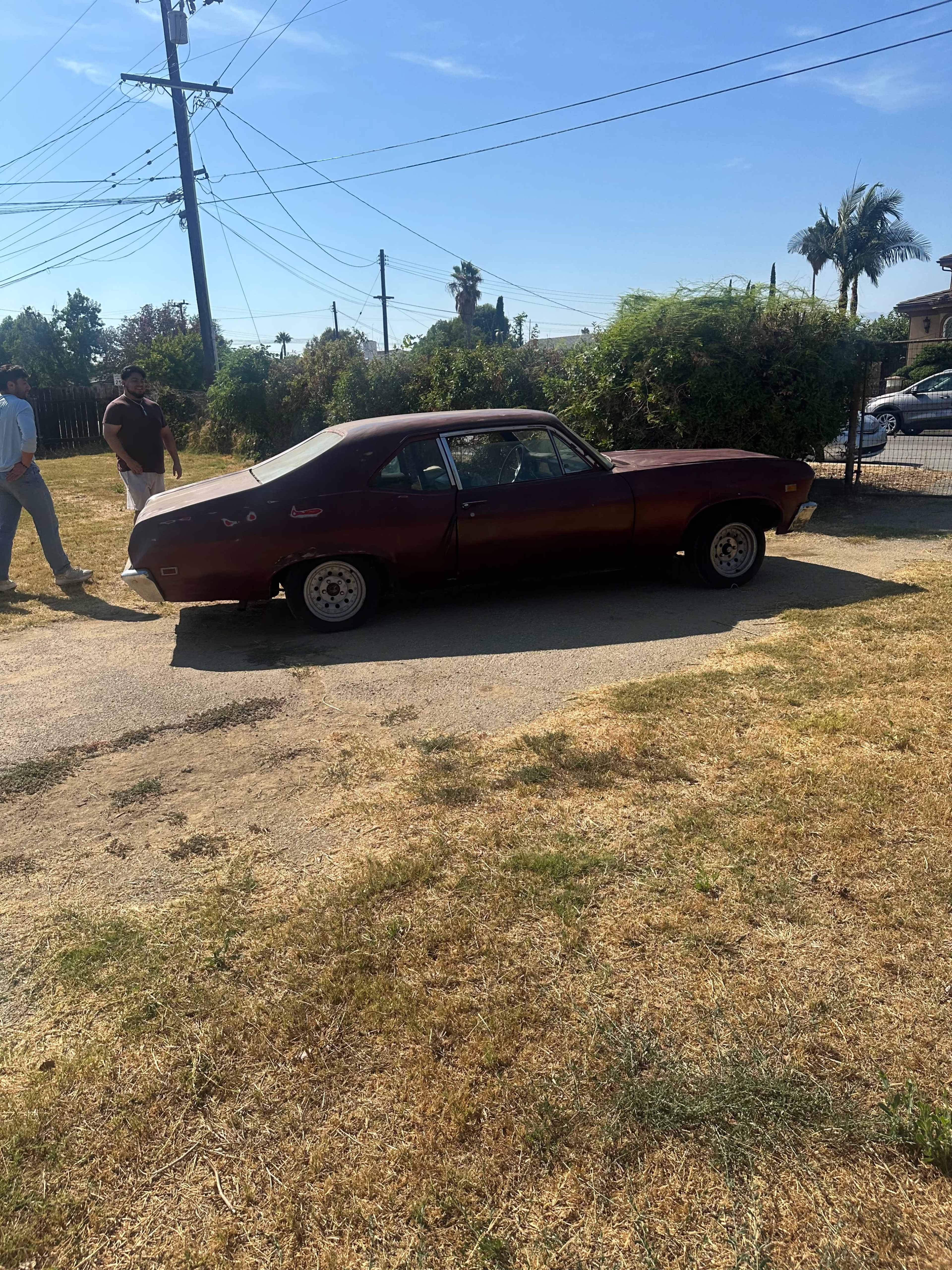 A classic red car is parked on a dirt path beside two people walking in a sunny outdoor setting.