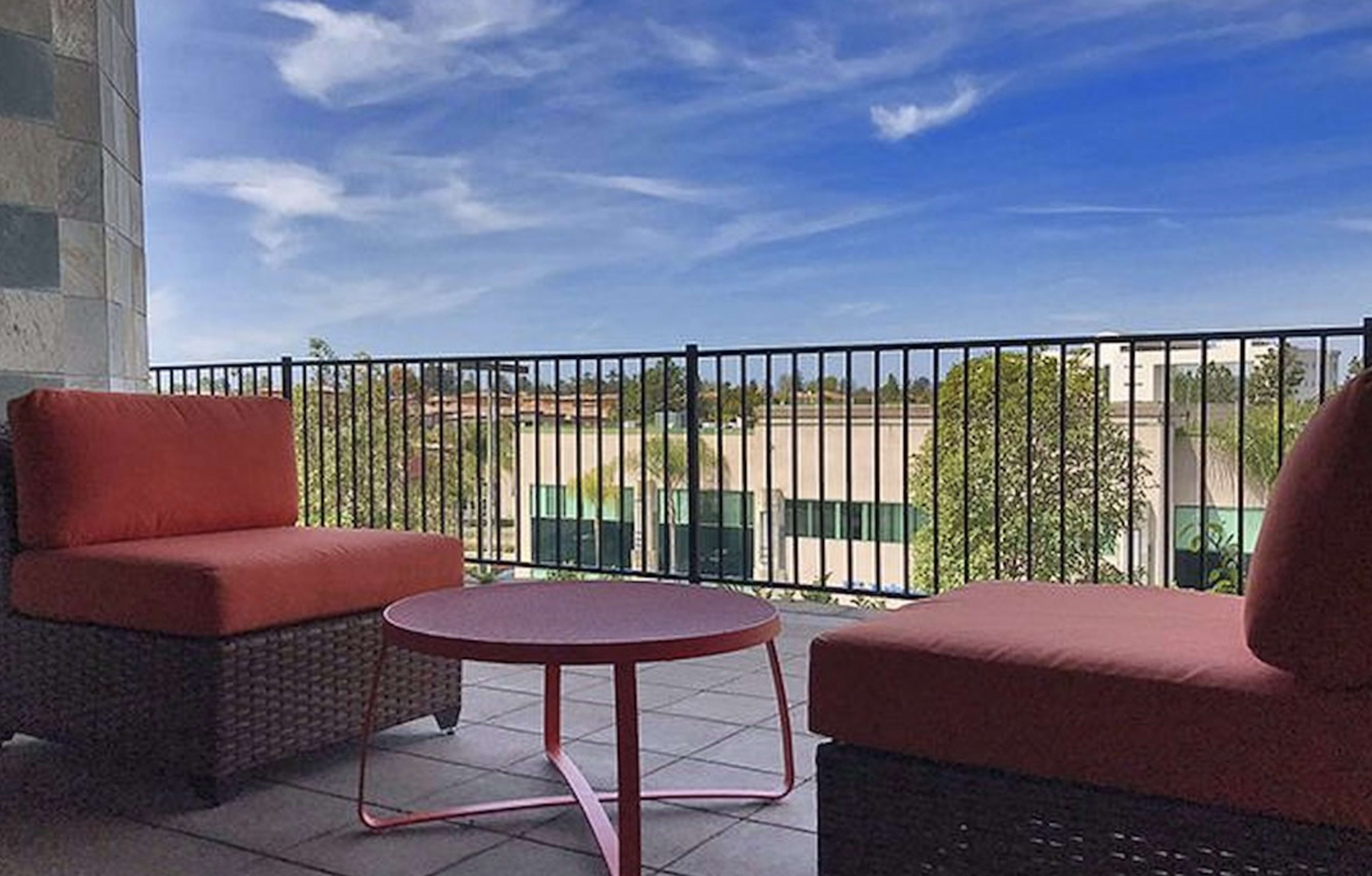 A balcony features two orange cushioned chairs and a round table, overlooking a view with trees and a blue sky.