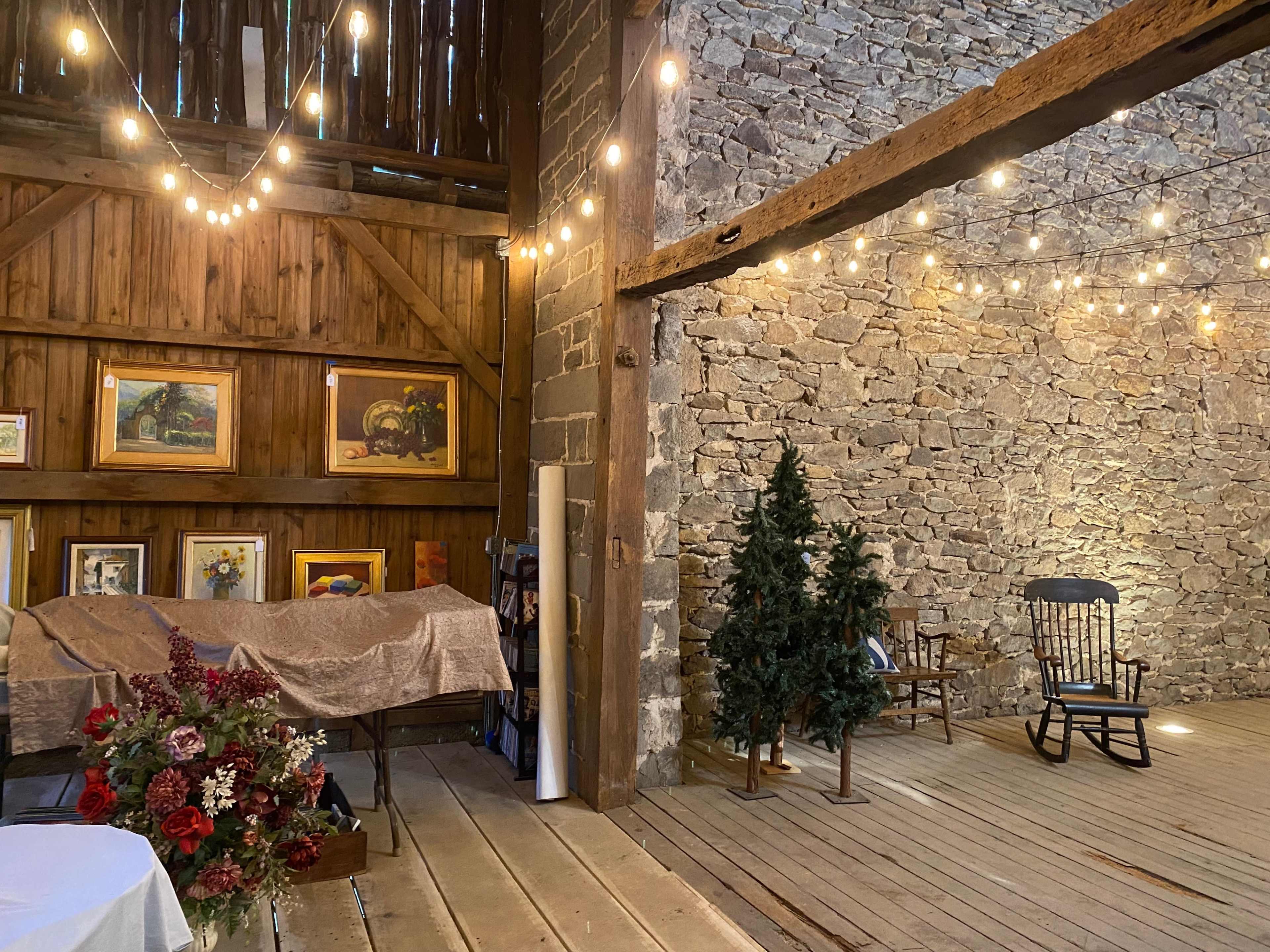 The interior of a rustic barn with stone walls, wooden beams, decorative string lights, and a small Christmas tree beside a rocking chair.