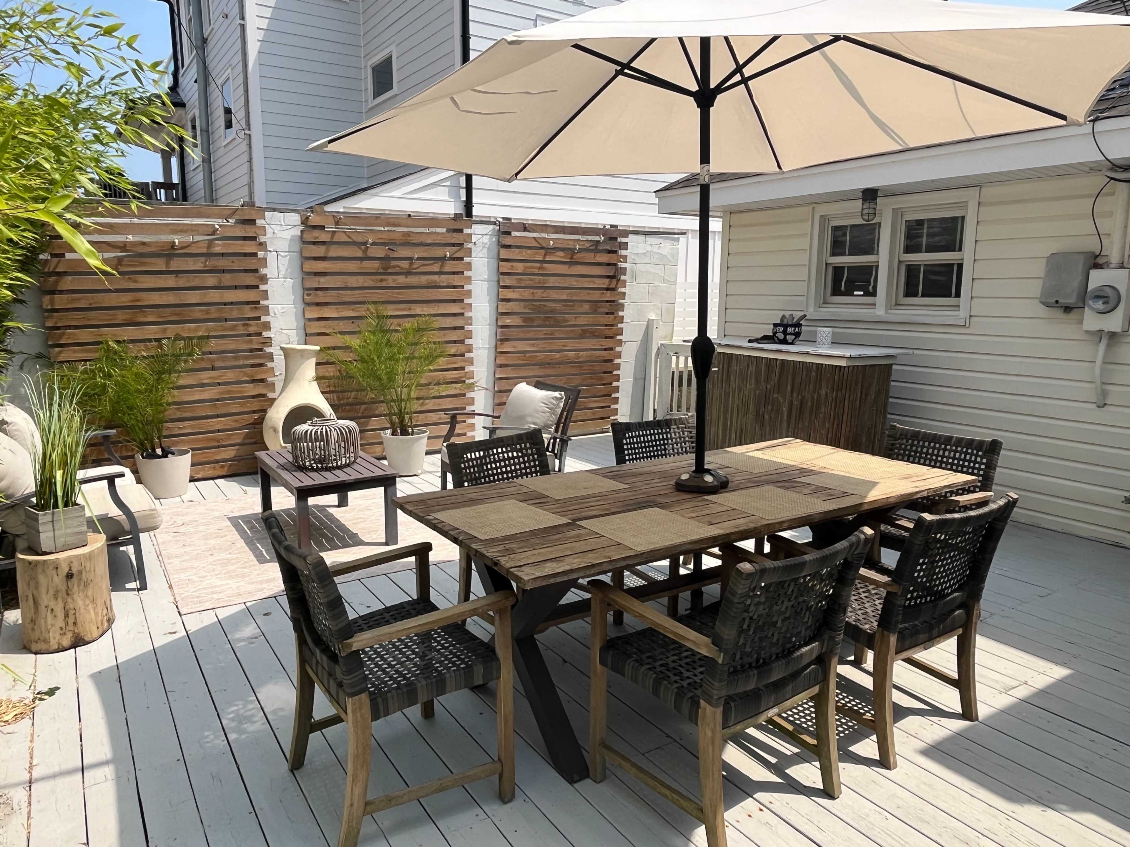 A backyard patio with a large wooden dining table, an umbrella for shade, and several chairs surrounded by potted plants and a bar area.