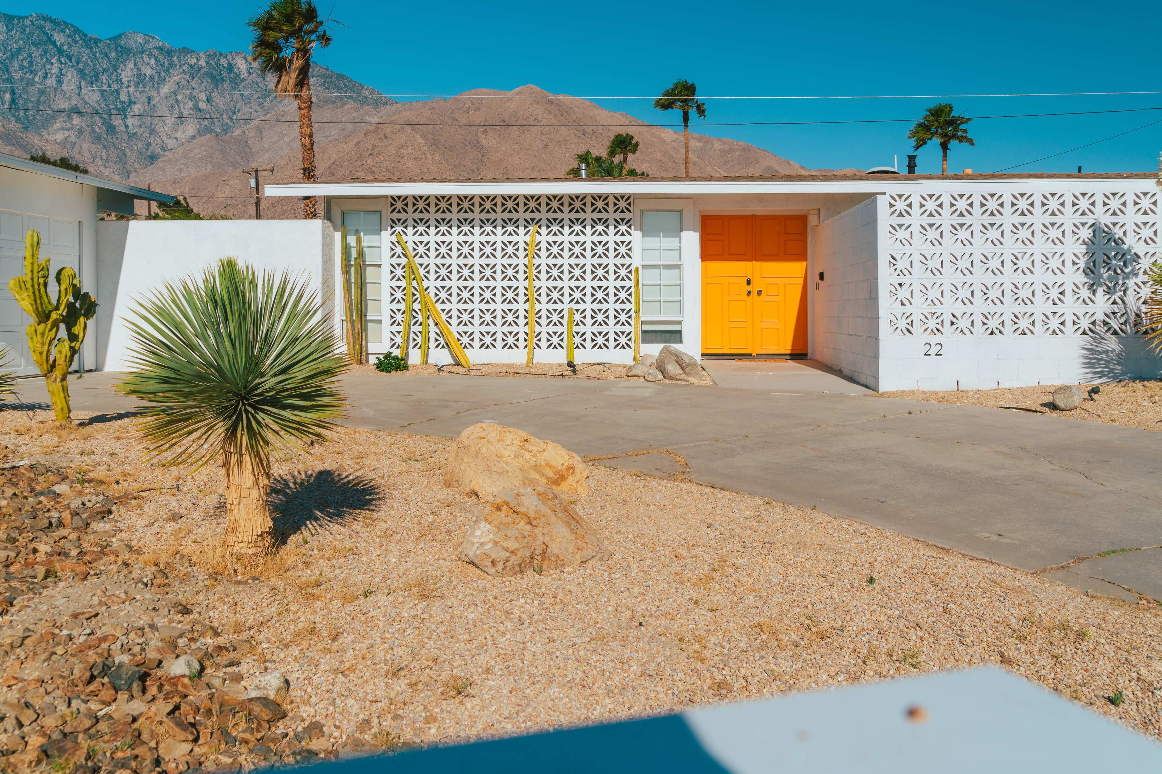 A single-story house with a bright orange door and a decorative wall stands in a desert landscape featuring palm trees and gravel.