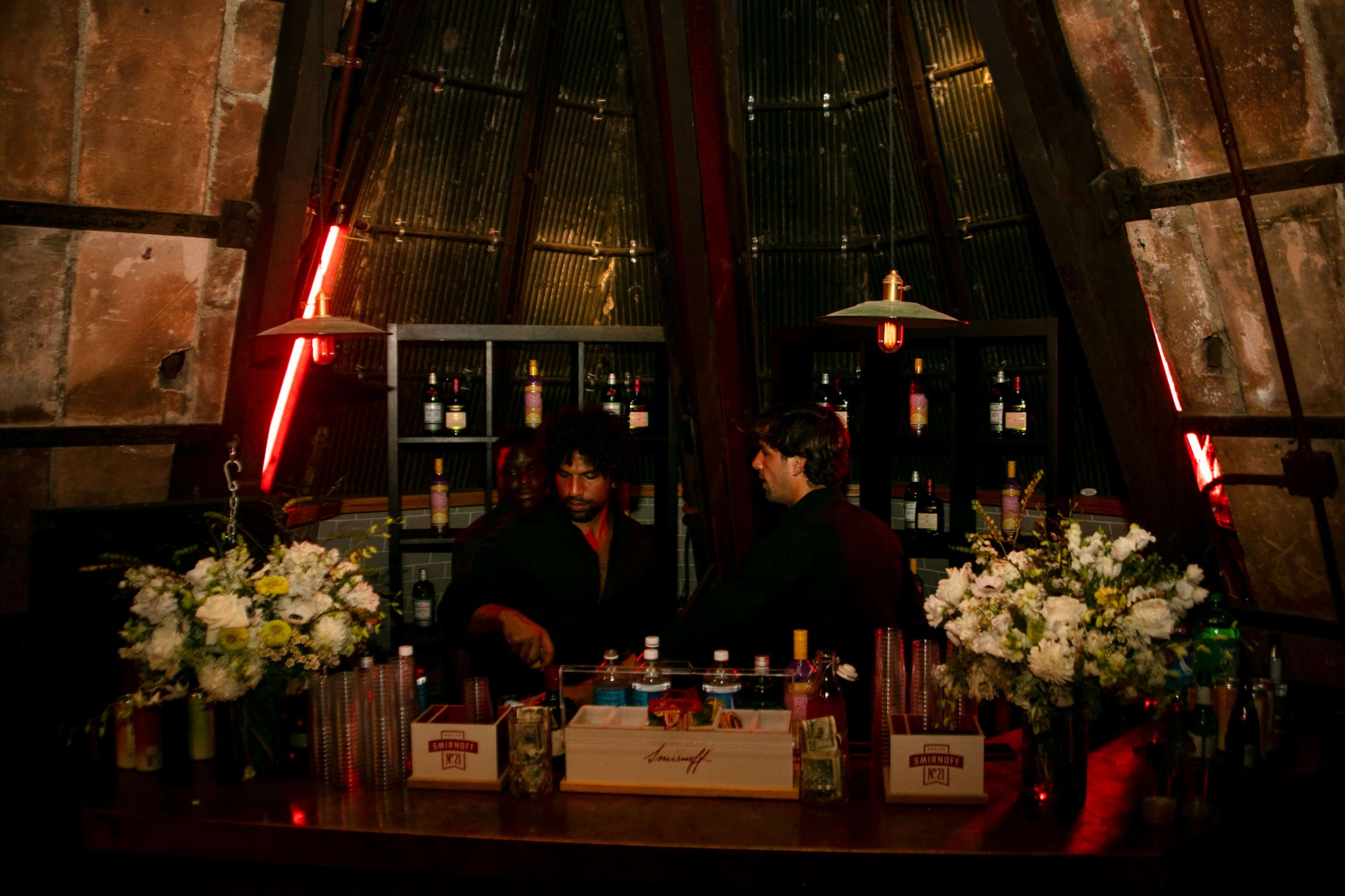 Two bartenders work behind a wooden bar adorned with flowers, while shelves filled with bottles line the walls of a dimly lit space with red neon lights.