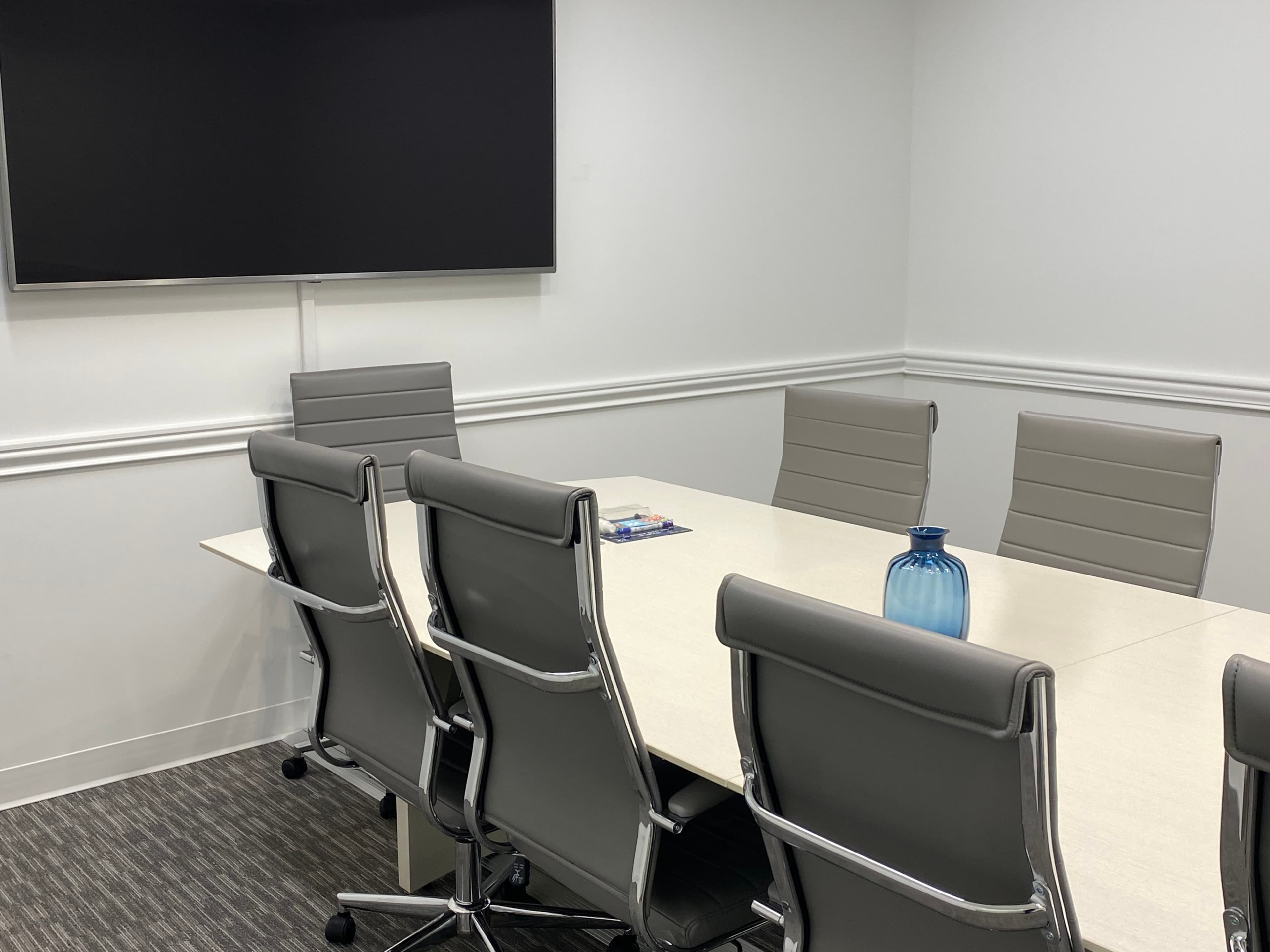 A conference room features a long table with six gray chairs, a blue vase, and a wall-mounted television.