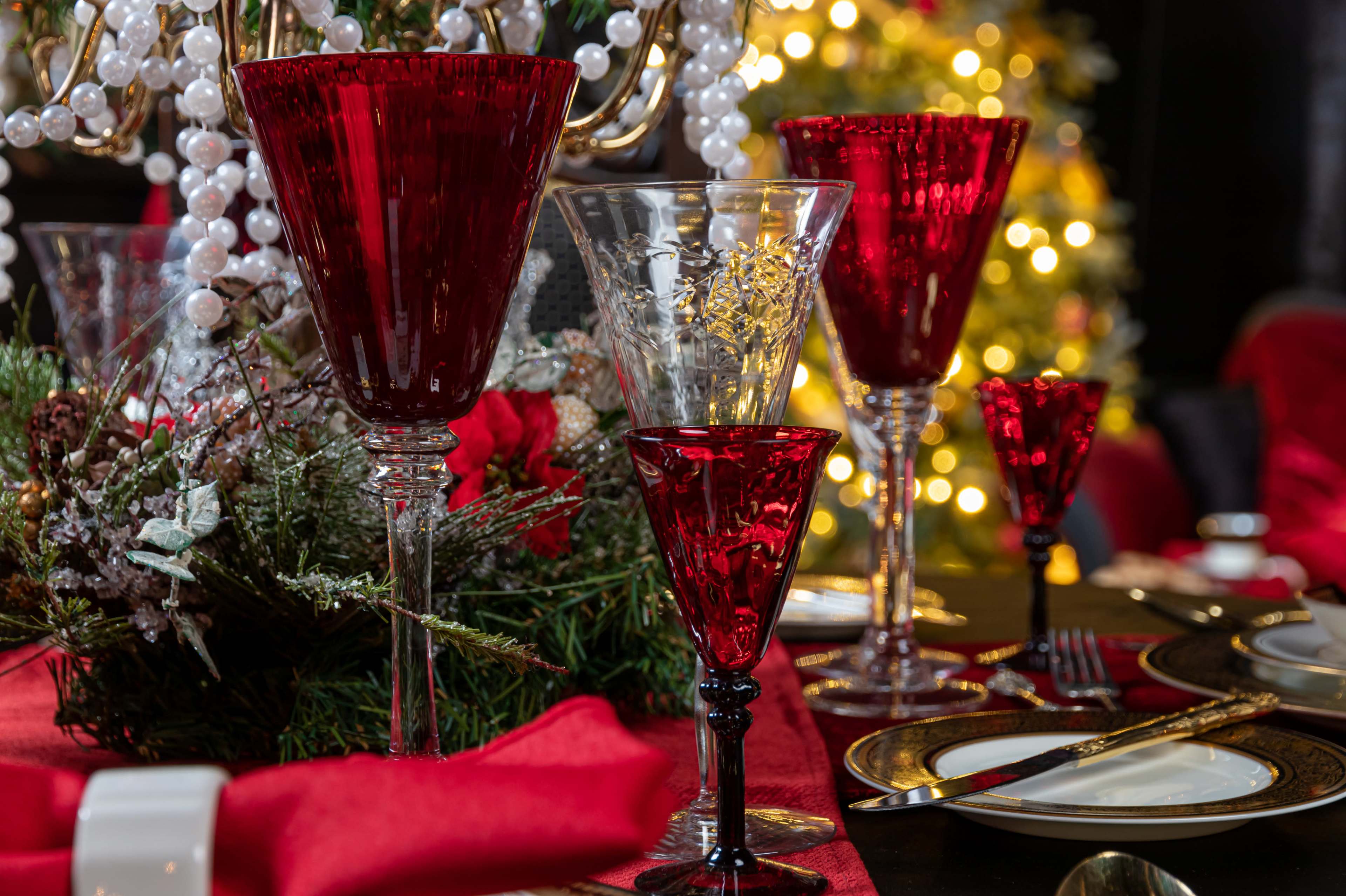 The image features an elegantly set dining table with red and clear glassware, decorated with greenery and festive elements, against a backdrop of a softly lit Christmas tree.
