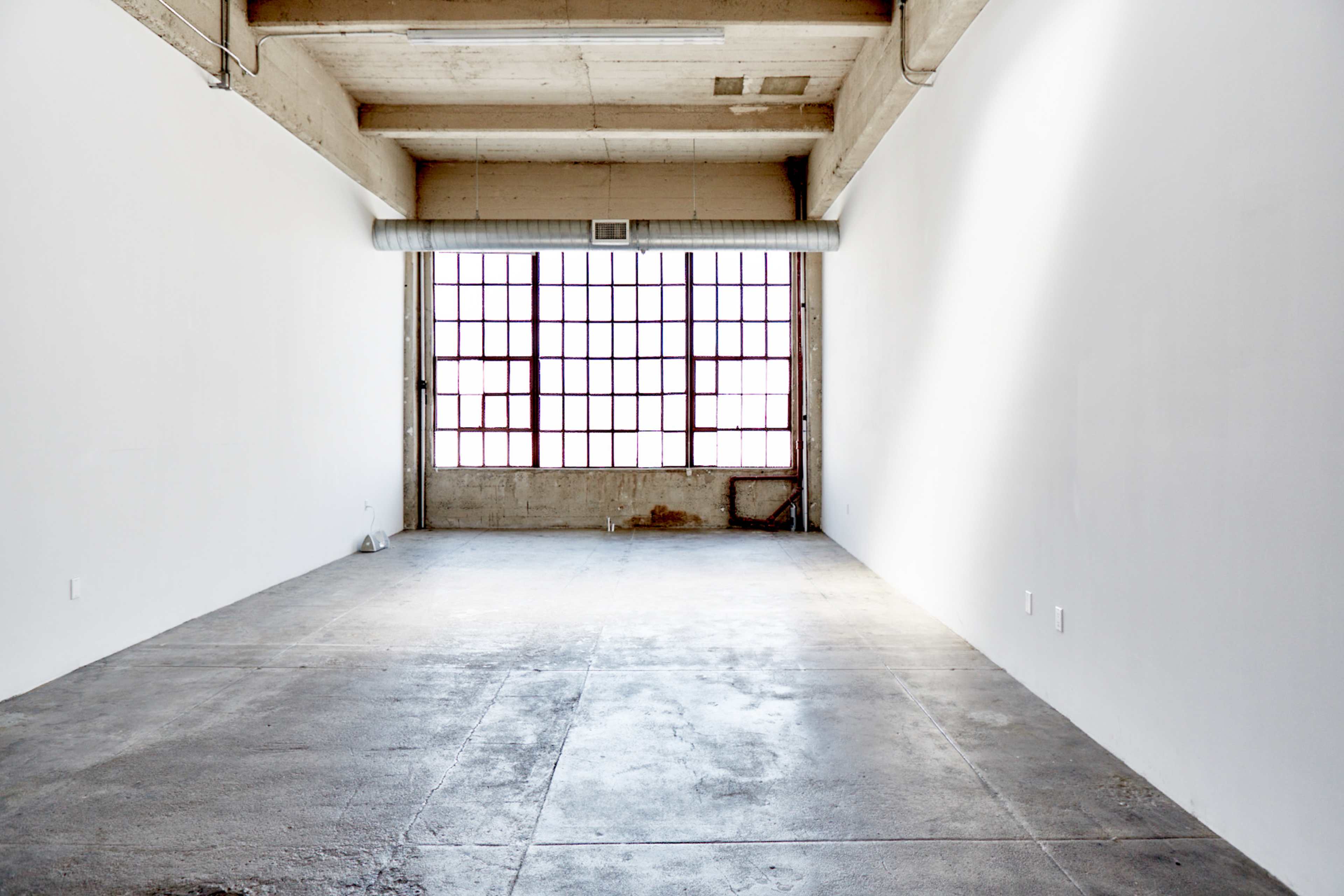 An empty room with concrete flooring and large windows along one wall, allowing natural light to enter.