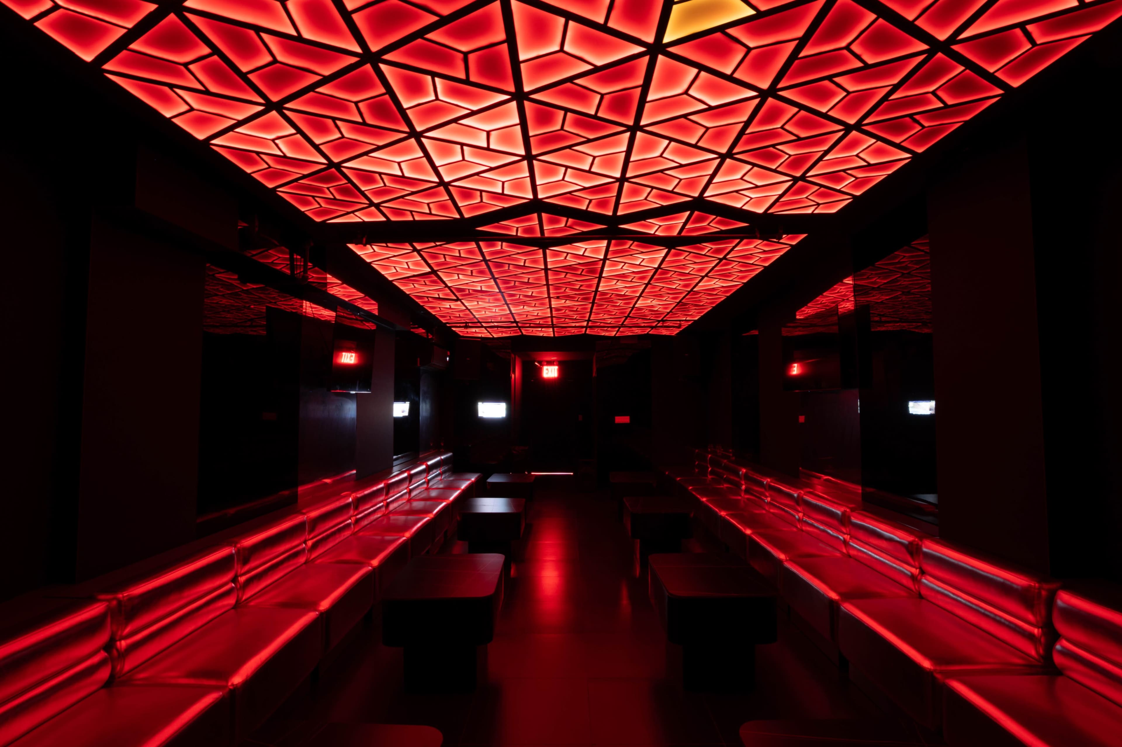 The image shows a dimly lit interior space with red geometric-patterned ceiling lighting and sleek black benches along the walls.