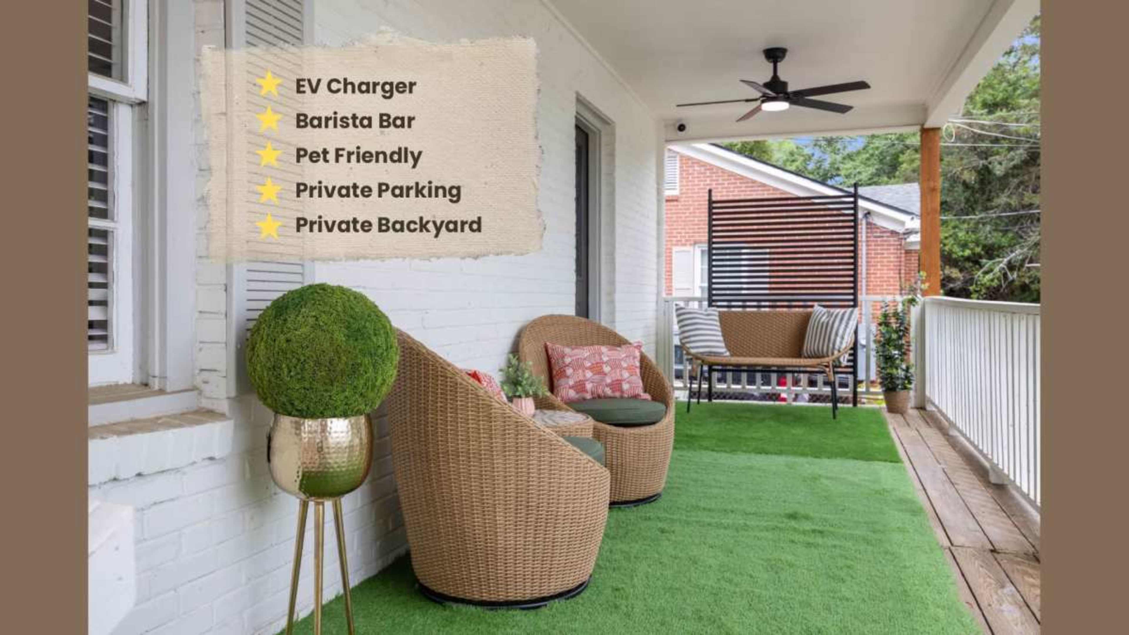 The image shows a porch with comfortable wicker seating, green plants, and decorative cushions, featuring a backdrop of a brick building and a shaded area.
