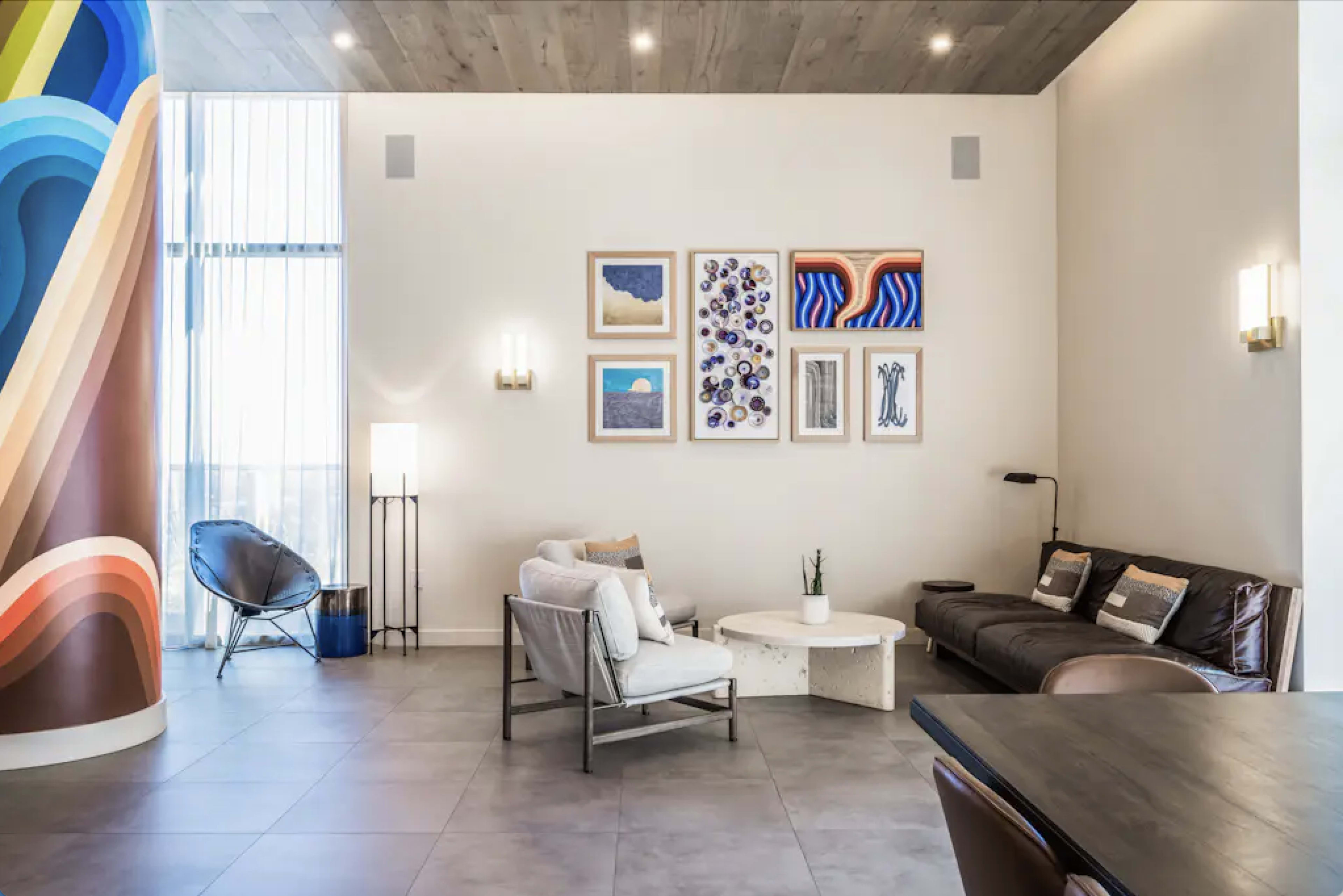 The image shows a modern living room with a mix of seating options, including a dark sofa, armchairs, and art displayed on the walls alongside a colorful mural.