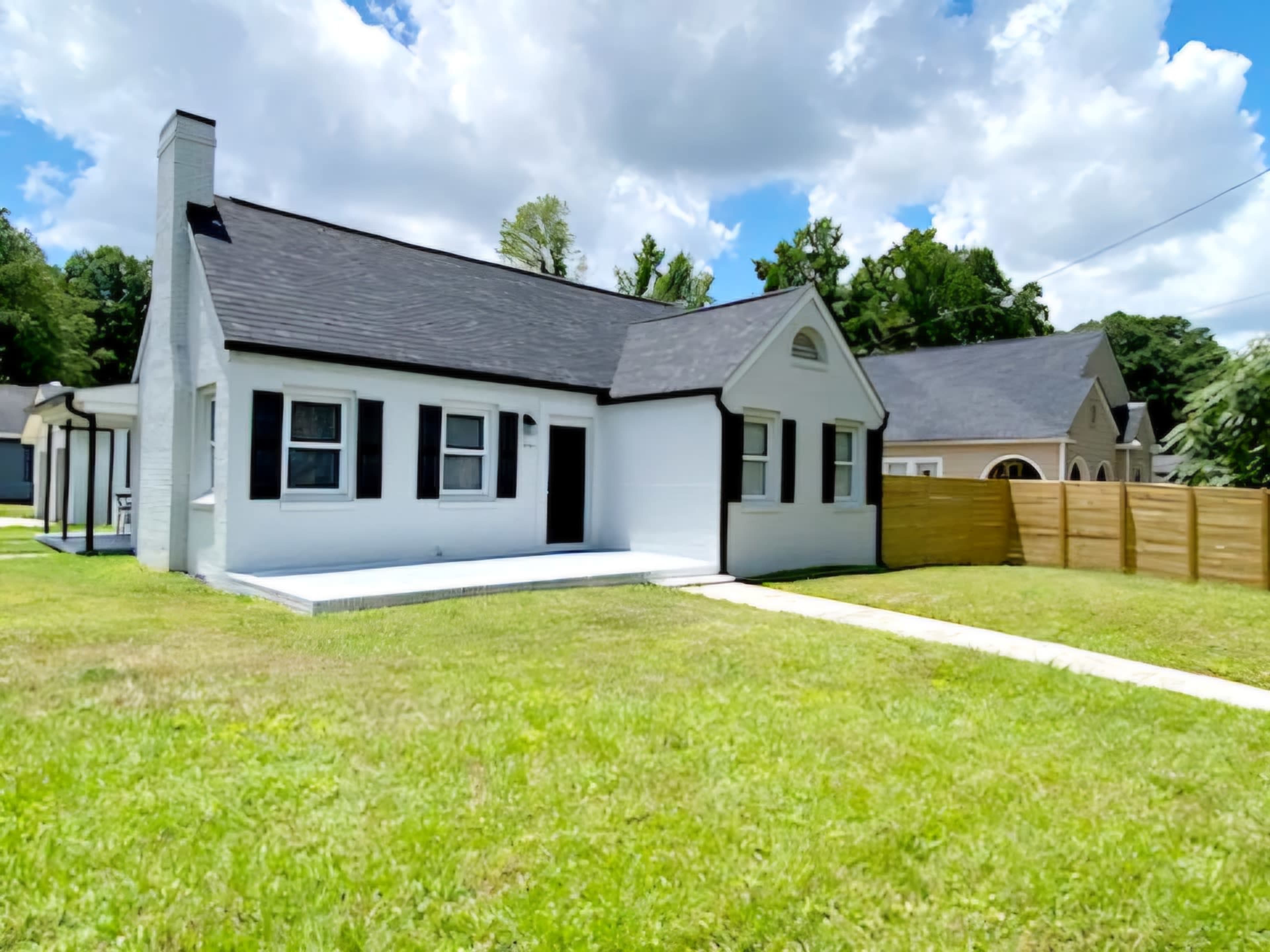 A white, single-story house with black trim and a front porch is set on a grassy lawn with a wooden fence in the background.
