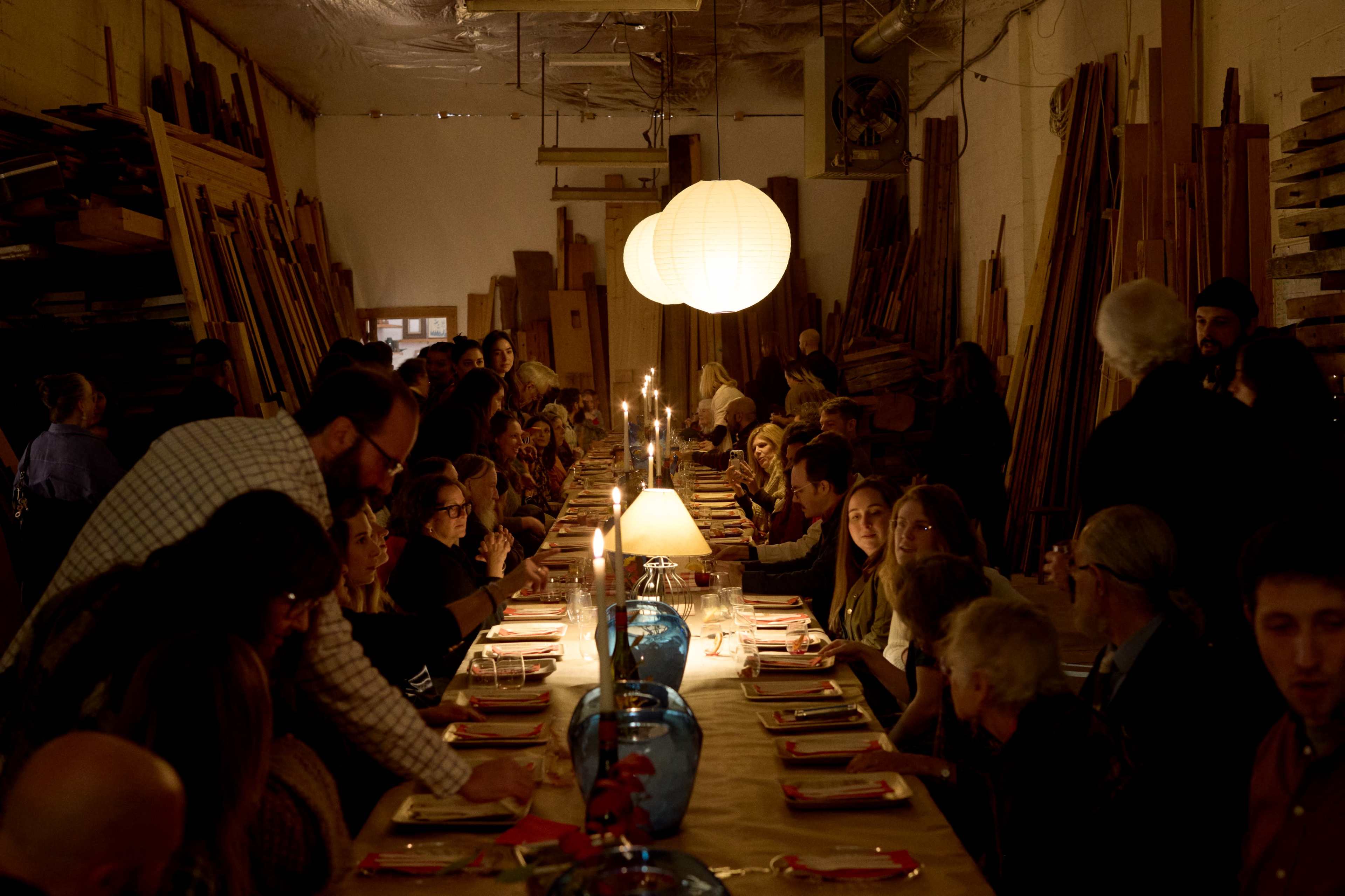 A large group of people is seated at a long dinner table illuminated by candles and pendant lights in a workshop filled with wooden materials.