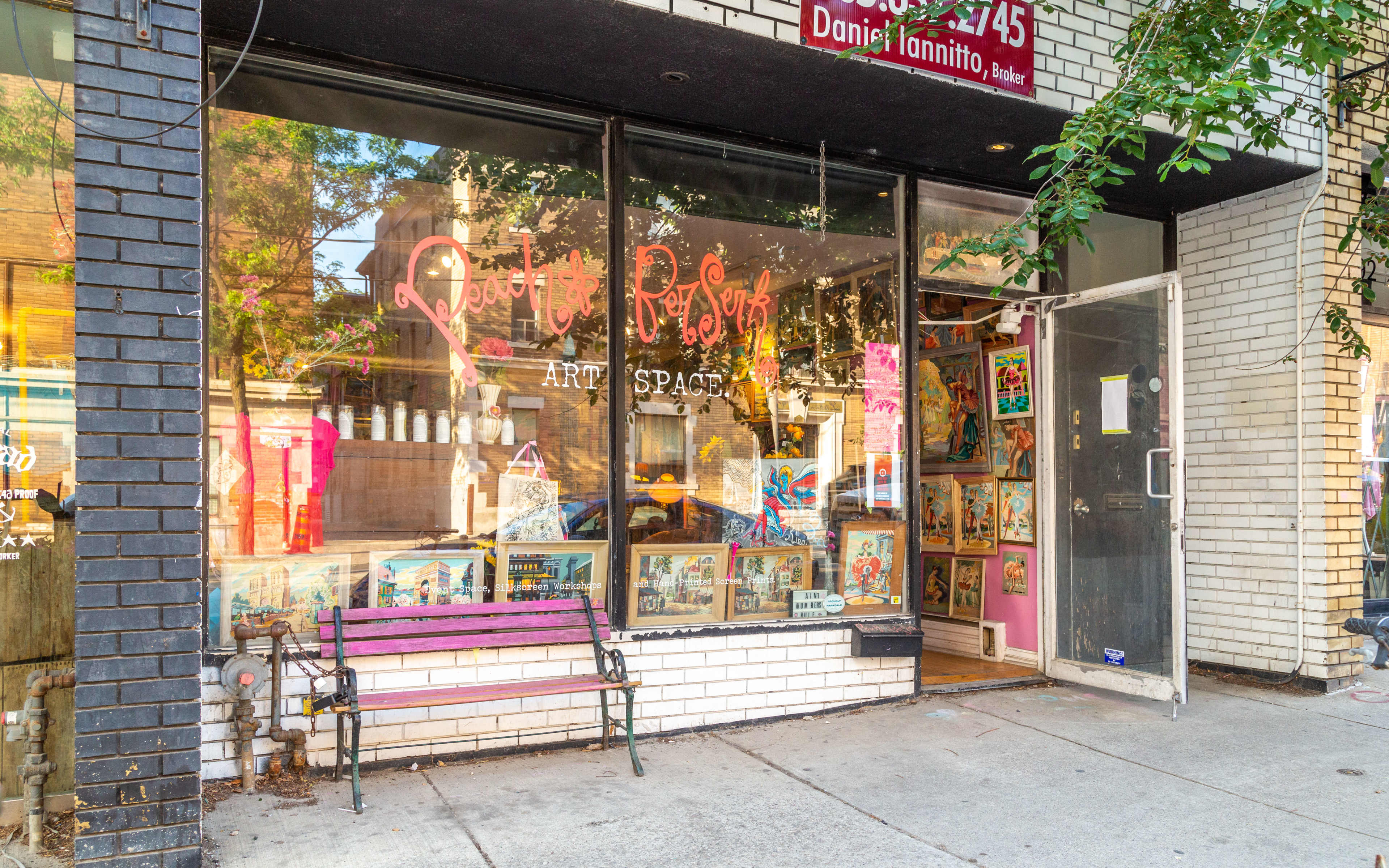 A storefront featuring large windows displays an array of framed artwork and a bench is positioned outside.