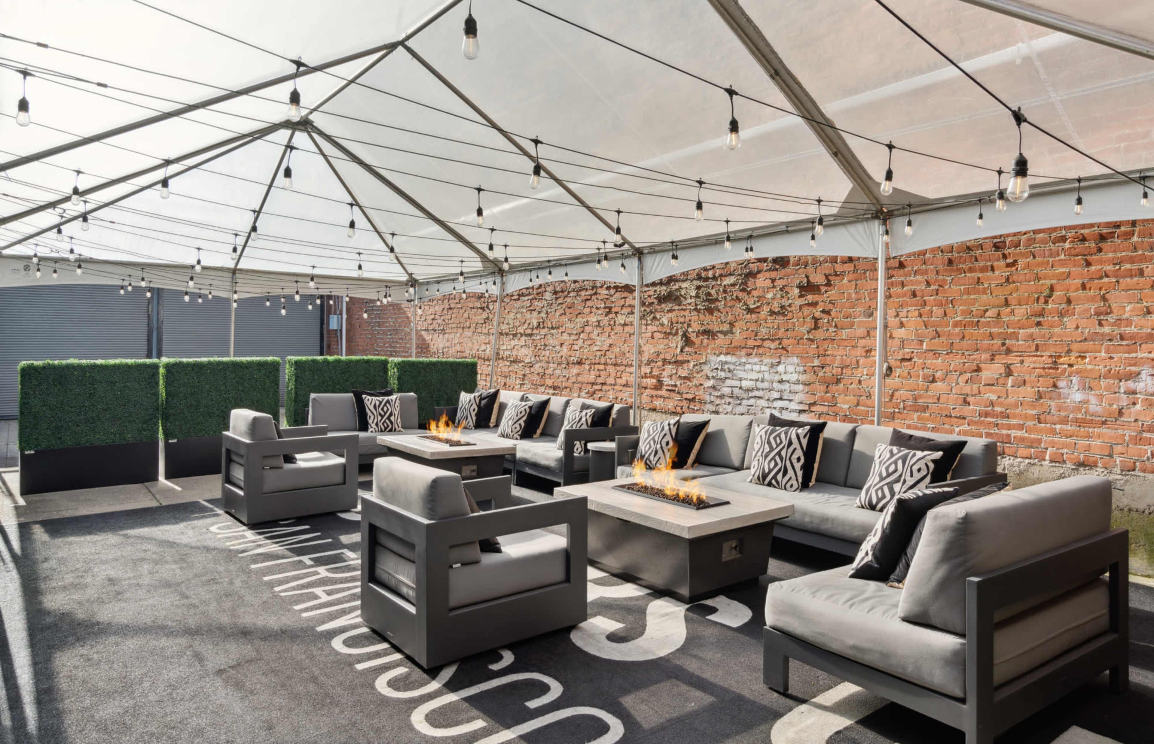 A covered outdoor lounge area features grey sofas arranged around a fire pit, with a backdrop of exposed brick walls and string lights above.