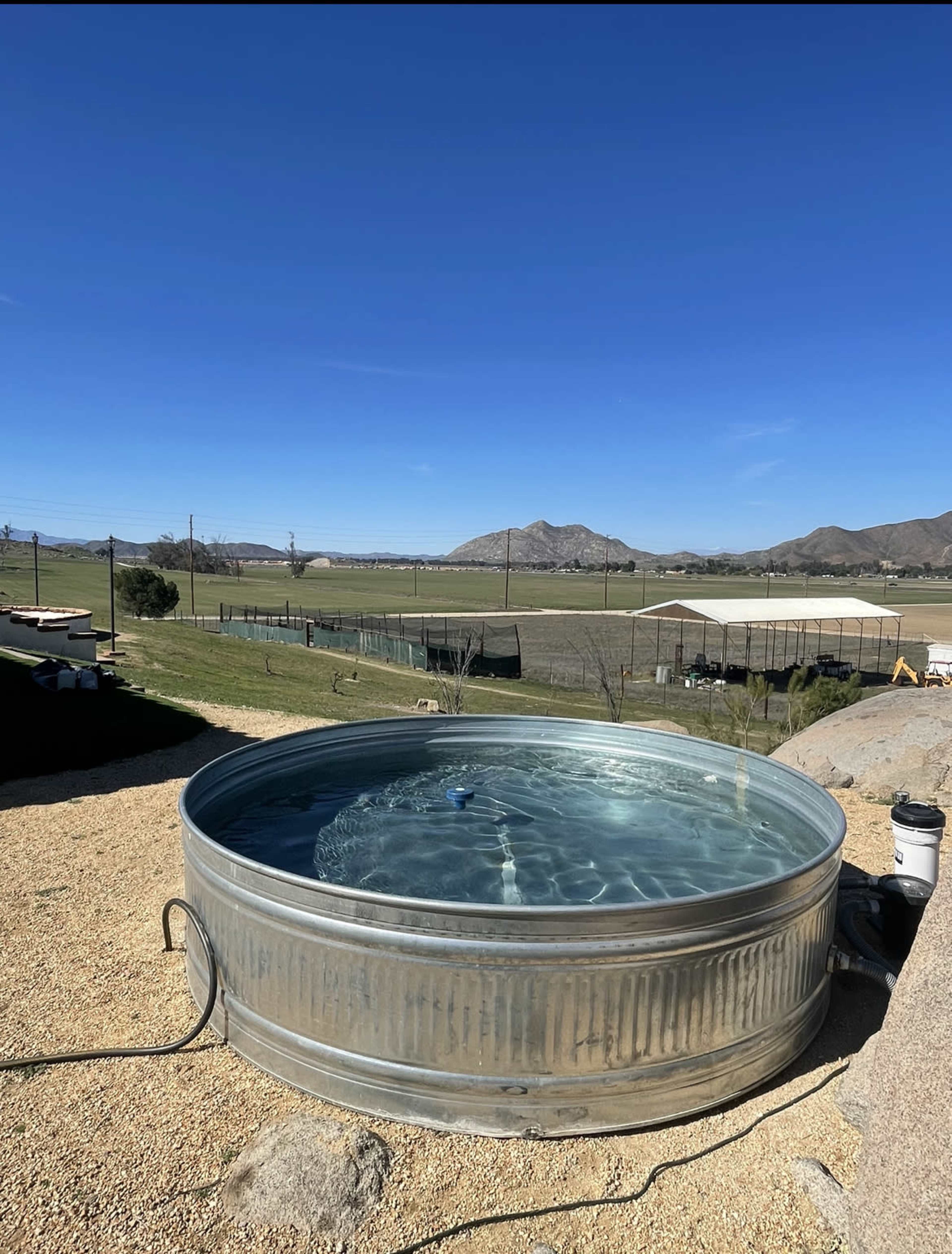 A round metal tub filled with water sits on a gravel area, overlooking a vast open field and mountains under a clear blue sky.