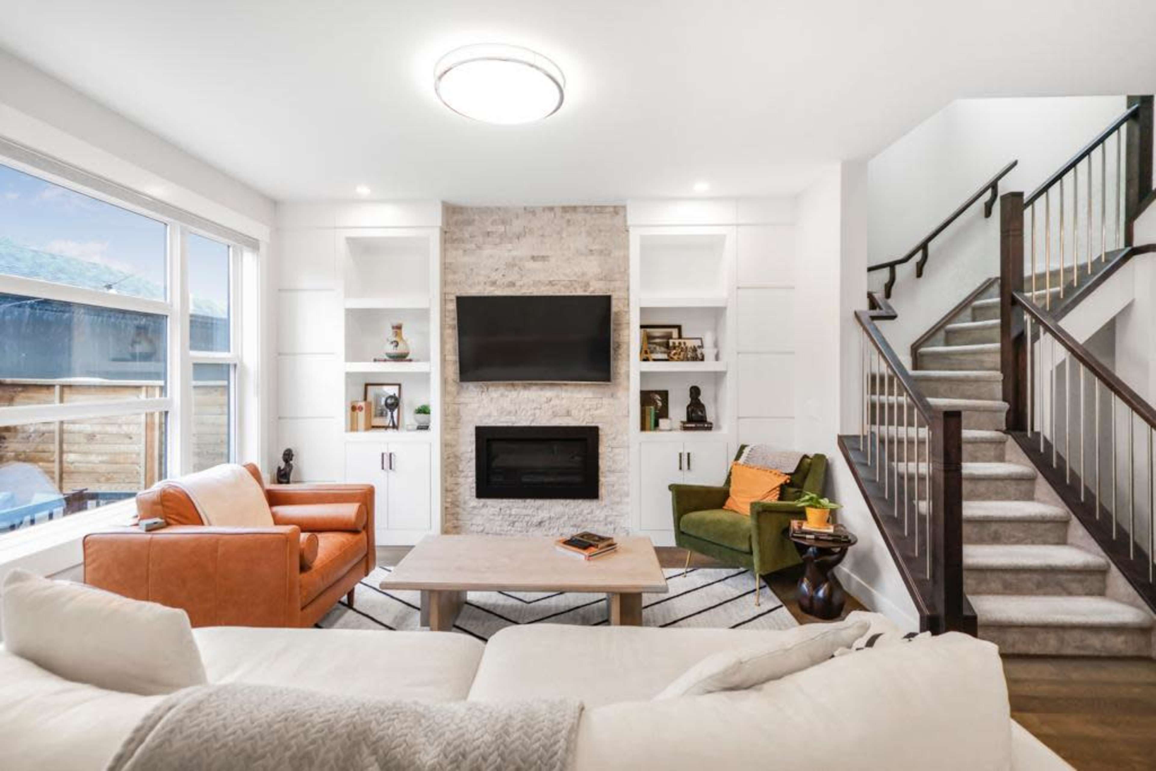 The image shows a modern living room featuring a fireplace with a TV above it, flanked by built-in shelves, and a staircase in the background.
