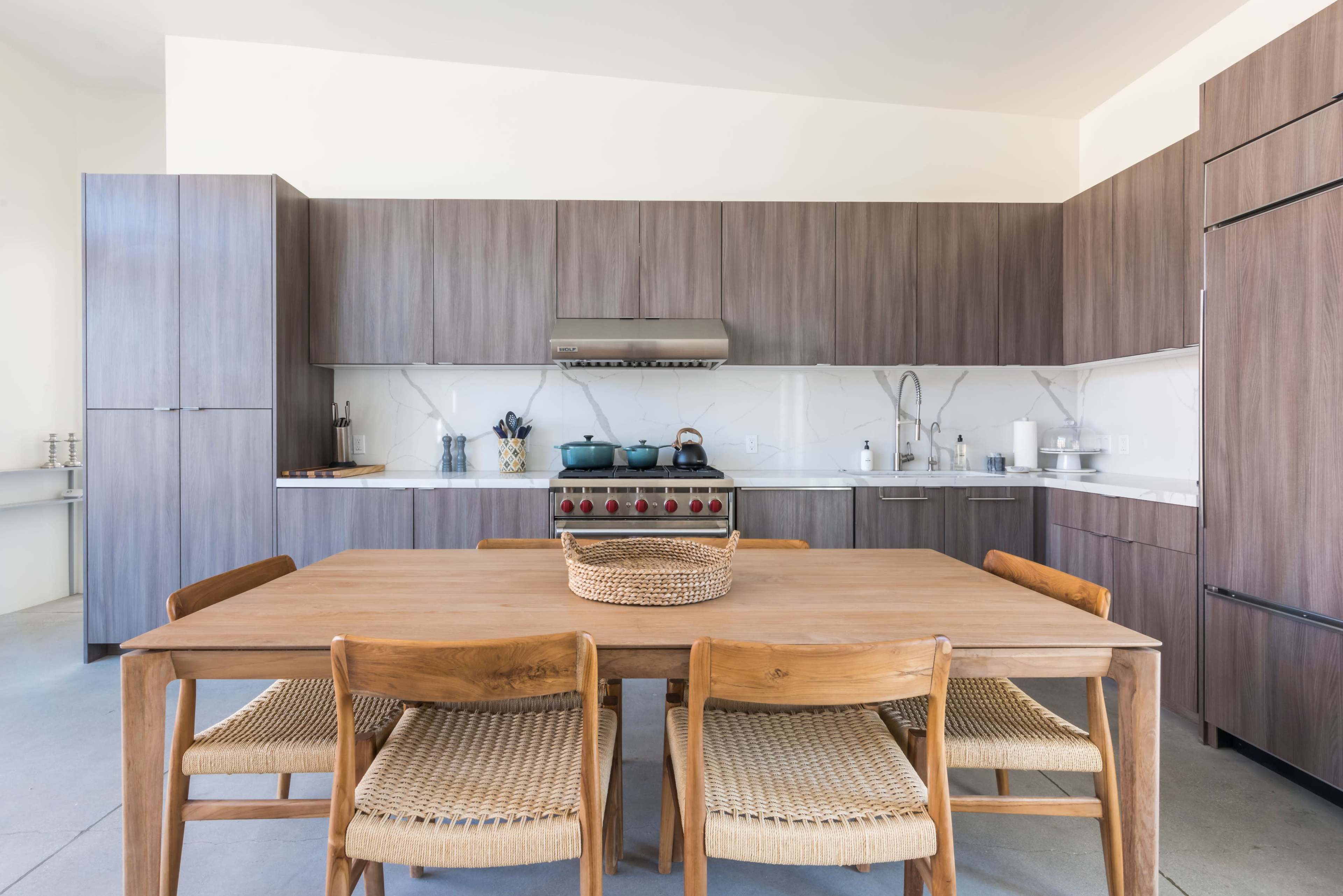 A modern kitchen features wooden cabinets, a central dining table with woven chairs, and stainless steel appliances against a light backdrop.