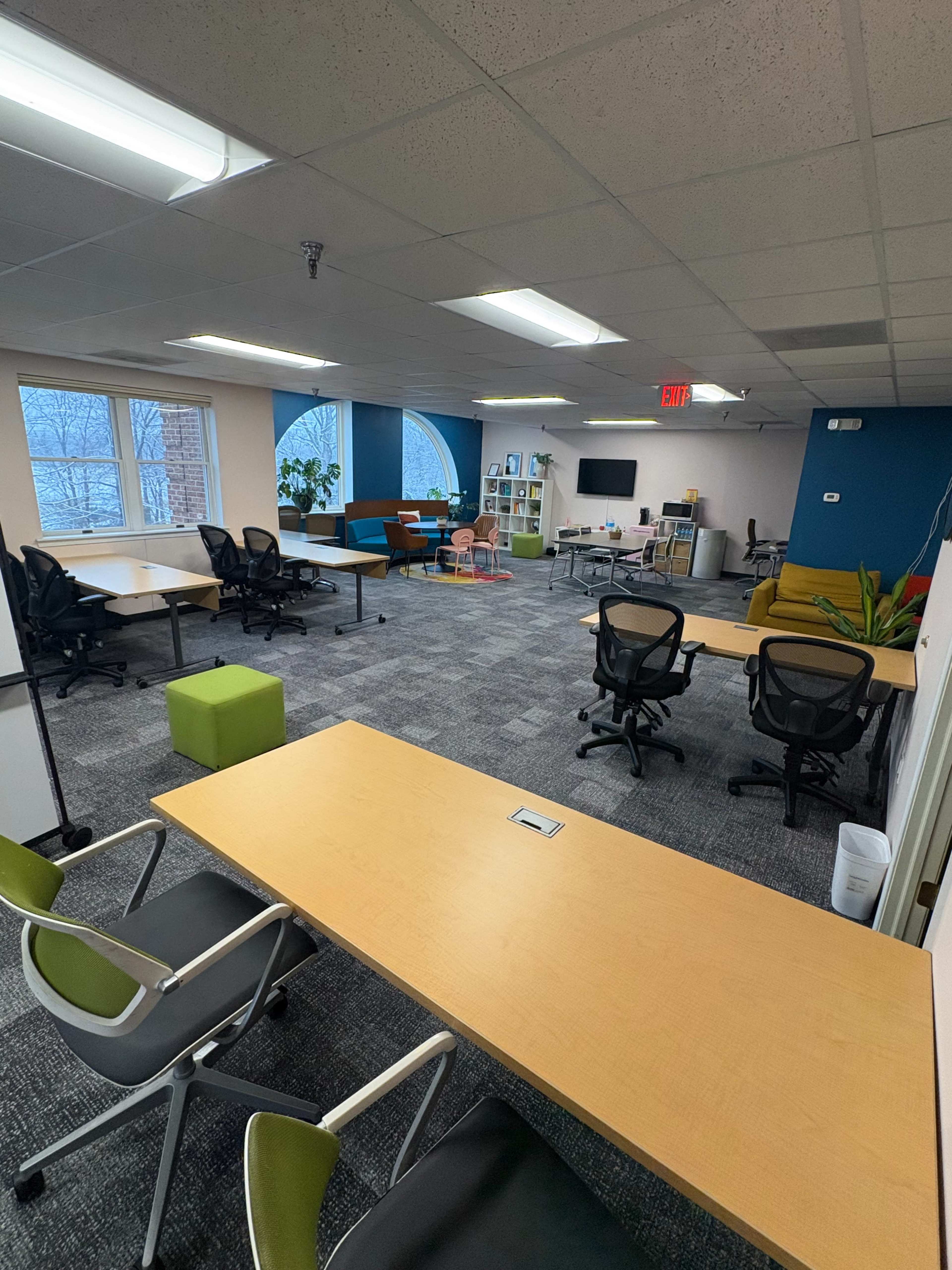 The image shows a spacious office area with several desks, chairs, and plants, featuring a blue accent wall and a large window.