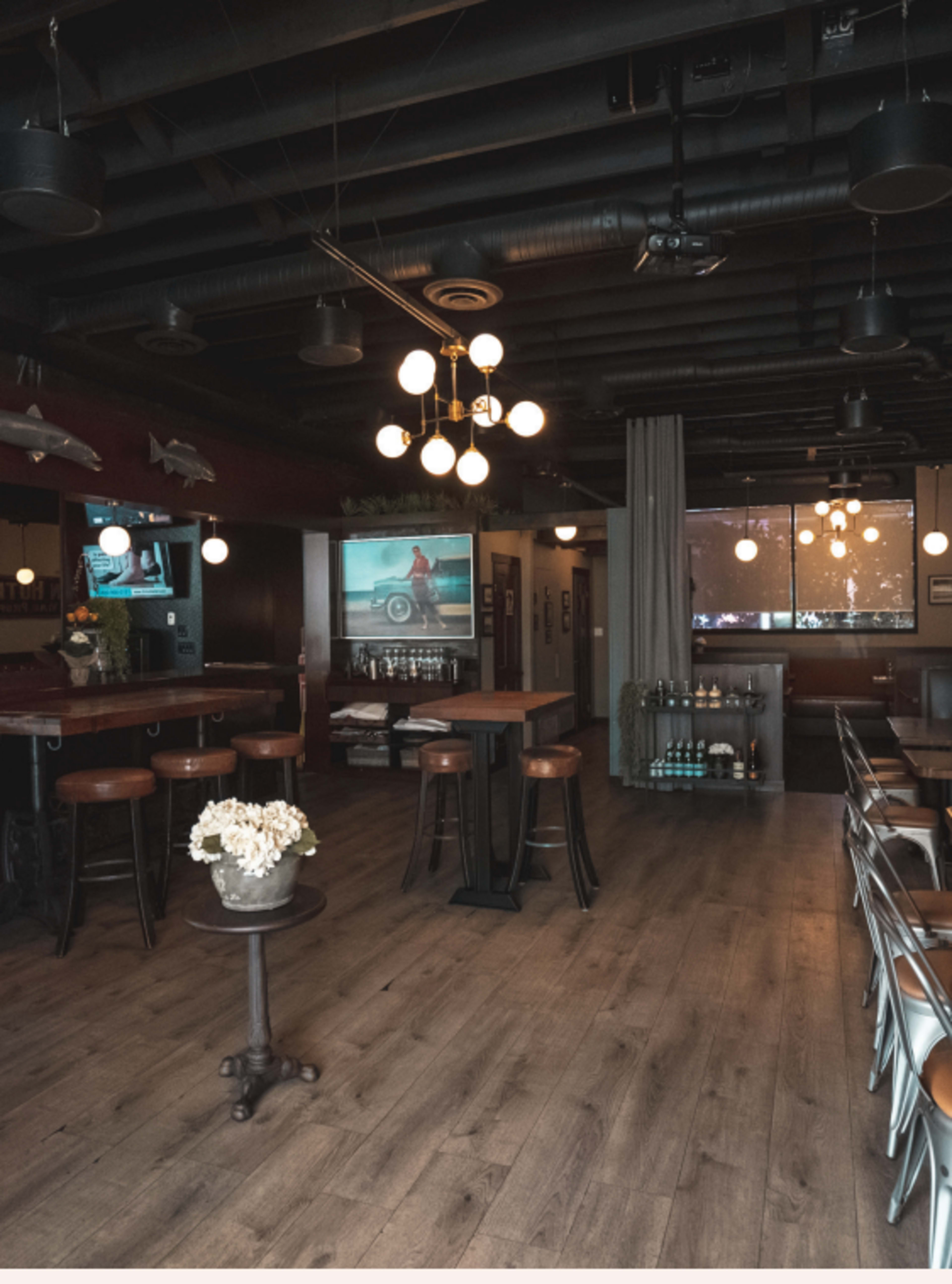 A modern restaurant interior with wooden floors, high tables, and ambient lighting, featuring a large screen displaying a video in the background.