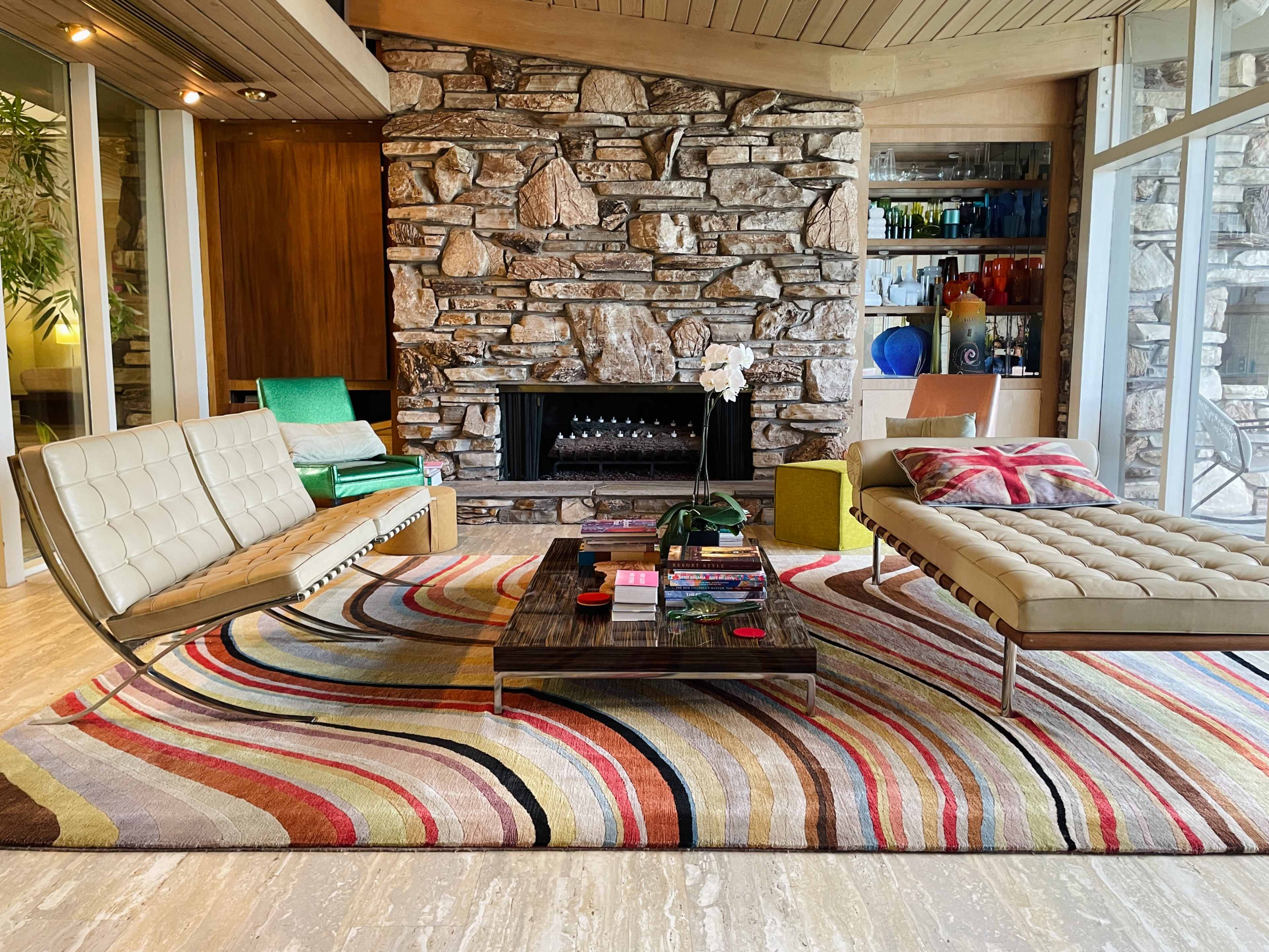 The image shows a modern living room featuring two distinct seating areas, a colorful area rug, a stone fireplace, and large windows allowing natural light.