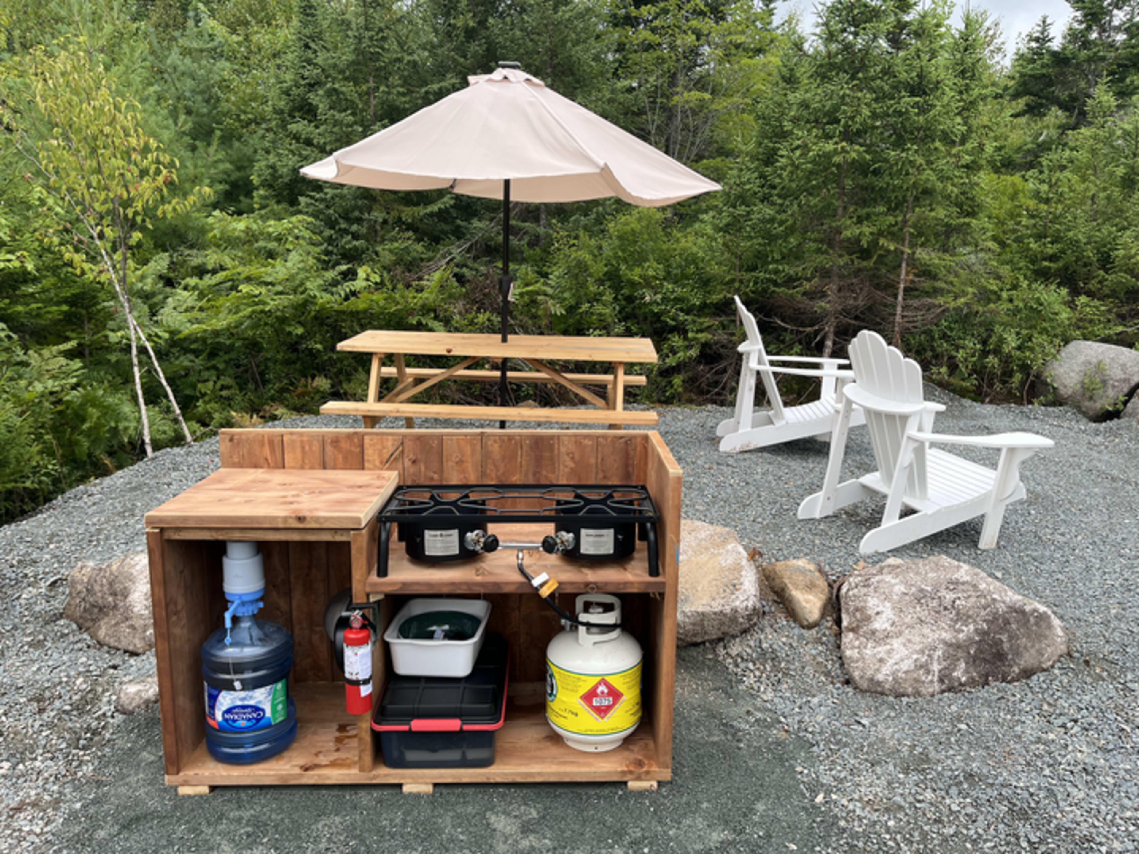 A wooden outdoor kitchen setup with a gas stove, water jug, fire extinguisher, and propane tank, surrounded by picnic tables and chairs beneath a large umbrella in a forested area.