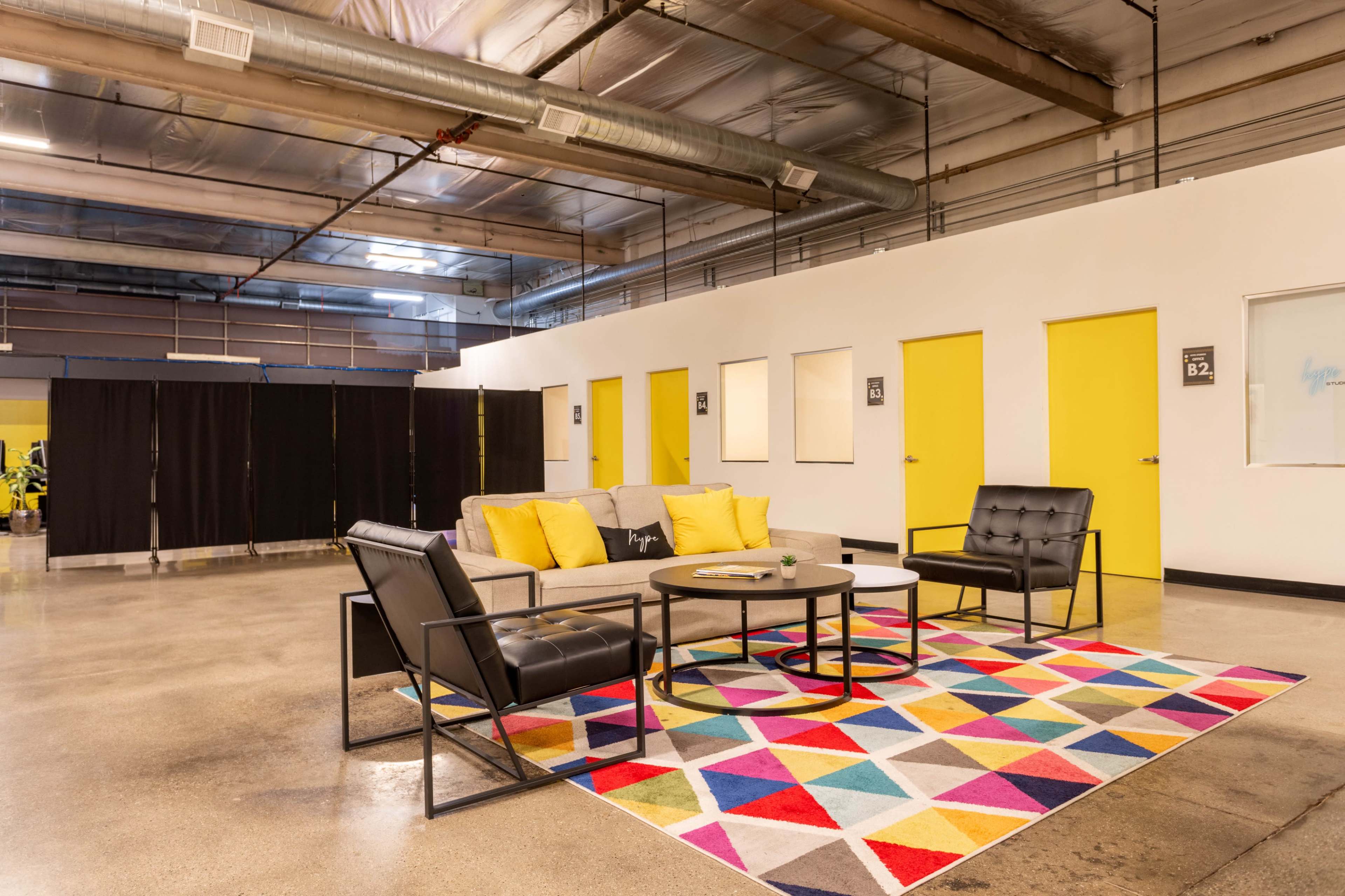 A spacious lounge area featuring a gray sofa with yellow cushions, two black chairs, a circular table, and a multicolored geometric rug on a polished concrete floor.
