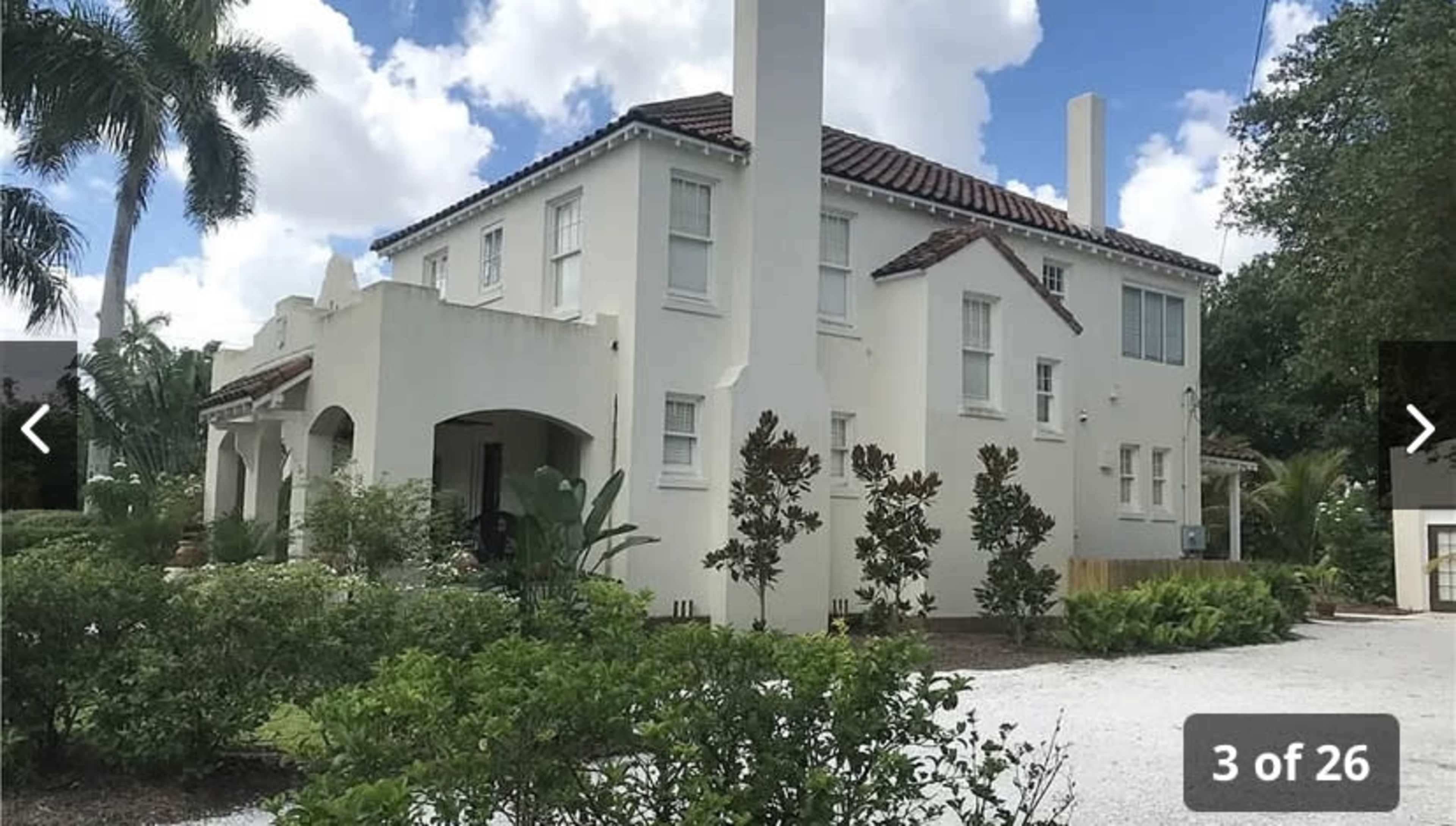 A two-story white house with a tile roof, surrounded by lush greenery and a gravel driveway.