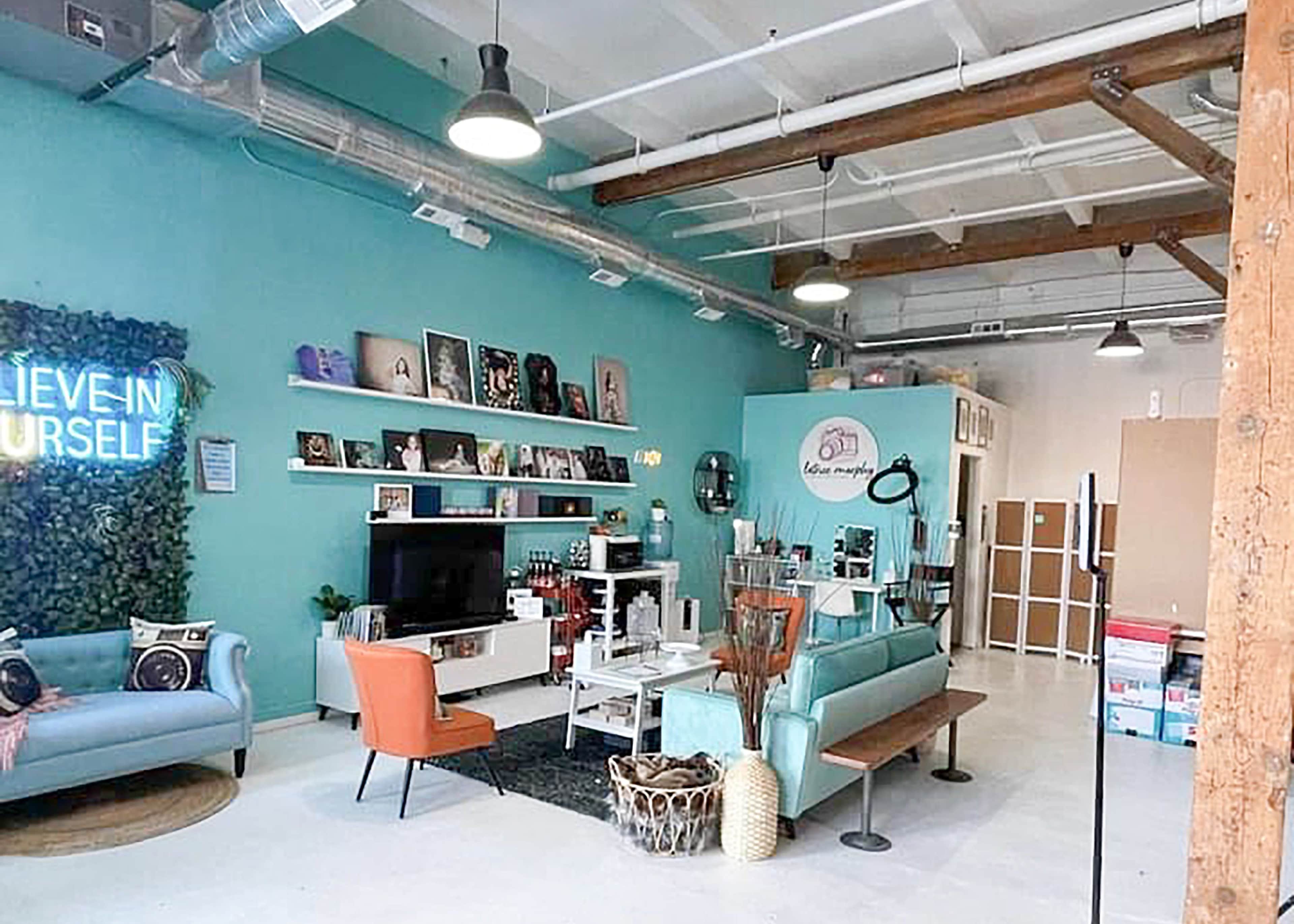 The image shows a brightly lit studio space with teal walls, modern furniture including a blue sofa and orange chair, and a display of artwork and decor items.