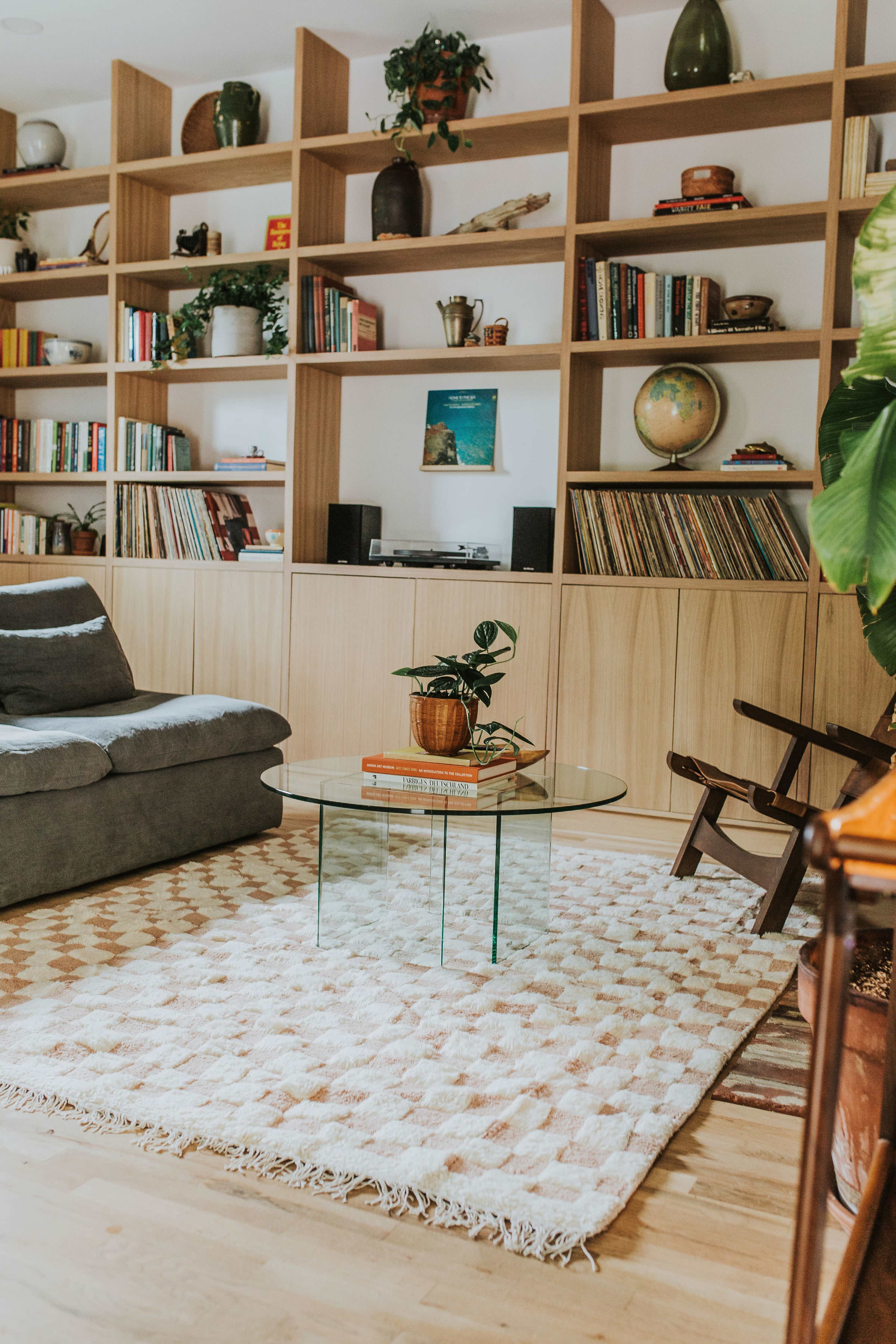 A cozy living room features a gray sofa, a glass coffee table with stacked books, and floor-to-ceiling shelves filled with plants and various decorative items.