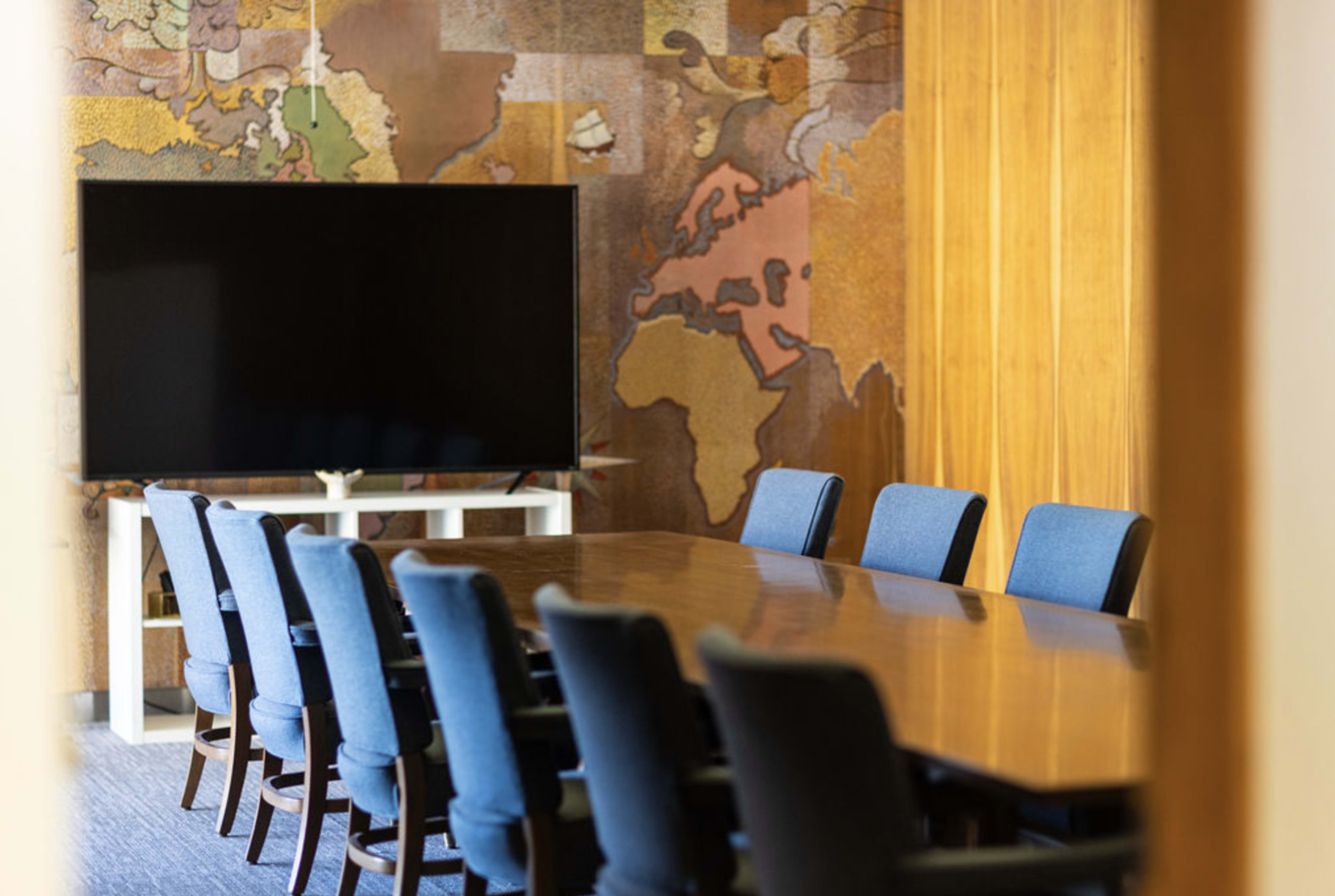 A conference room features a long wooden table surrounded by blue chairs, with a large TV screen and a world map mural on the wall.
