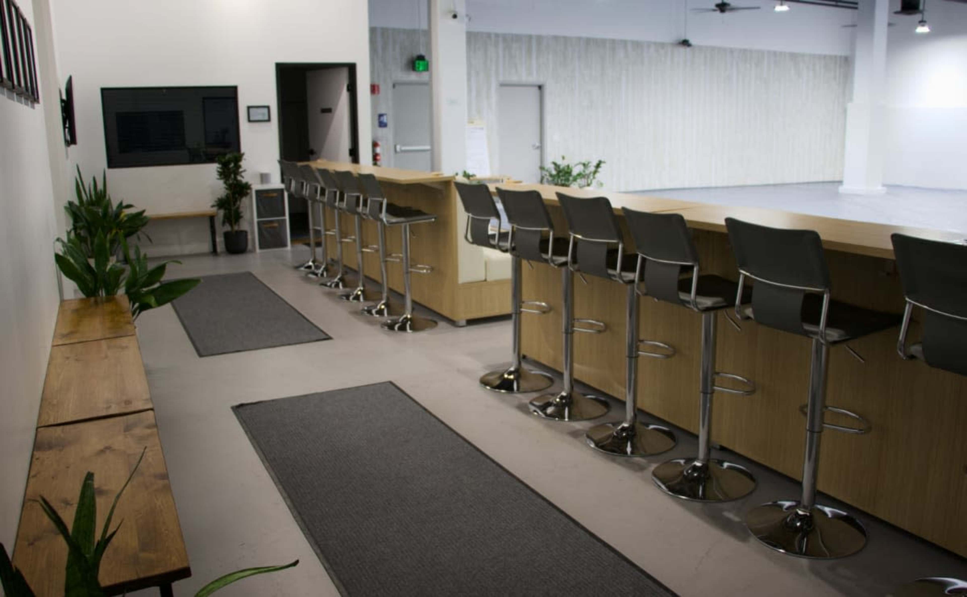 The image shows a modern interior featuring a long wooden counter with high chairs, a series of potted plants, and clean, minimalistic decor.