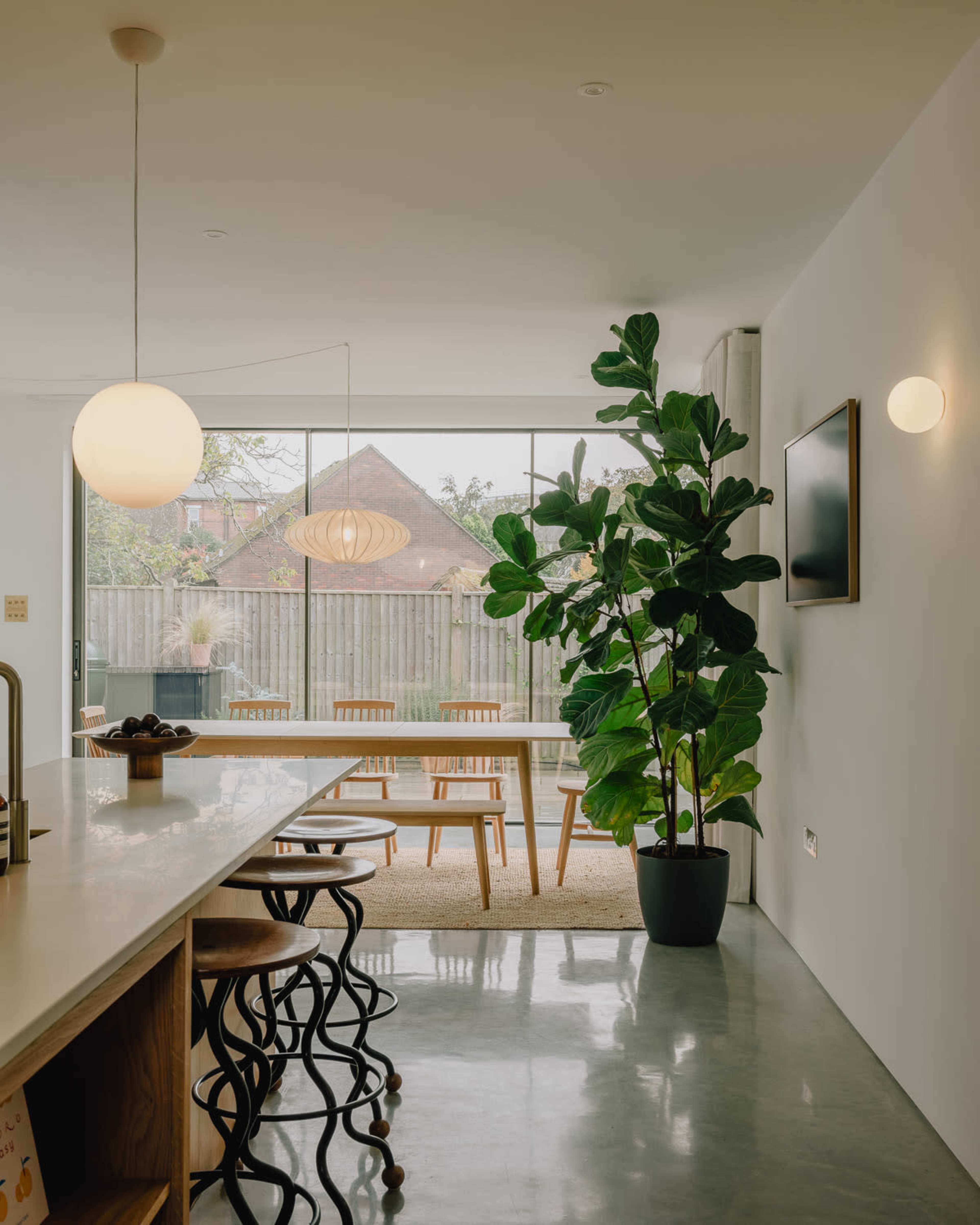 A modern, open-concept kitchen features a long white countertop, a large window with a view of a garden, and a prominent potted plant beside a dining area with wooden chairs.