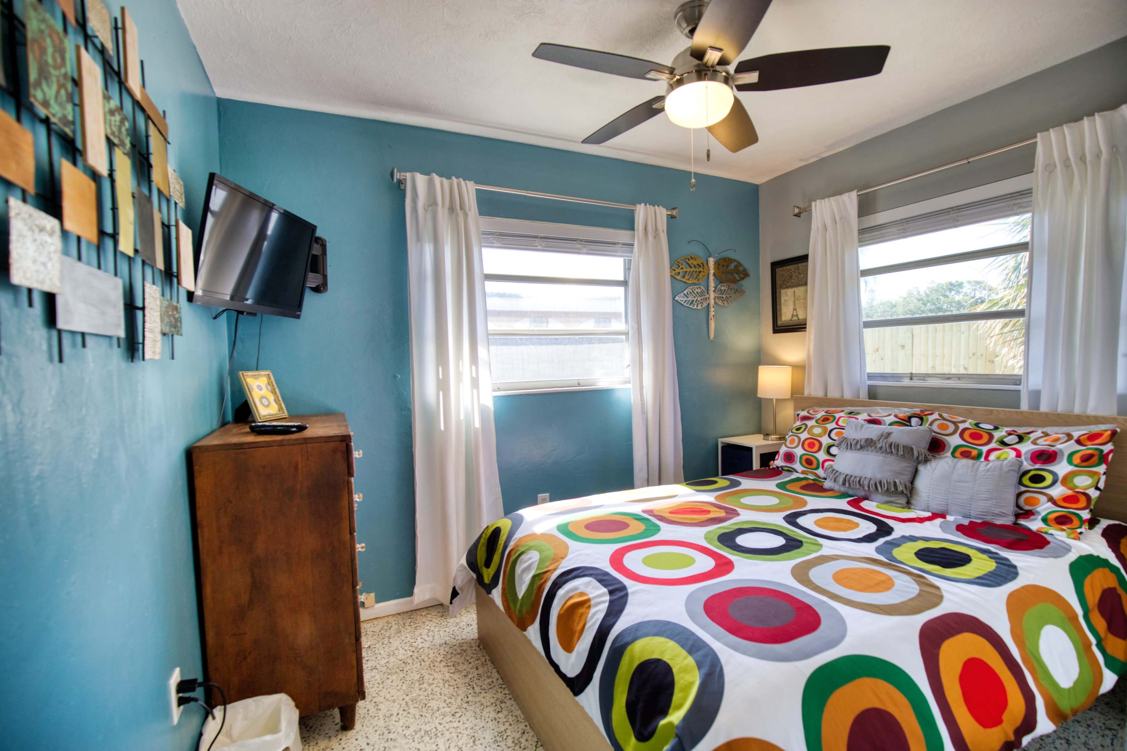 The image shows a bedroom with a colorful polka dot bedspread, a wooden dresser, a television mounted on the wall, and windows with white curtains.