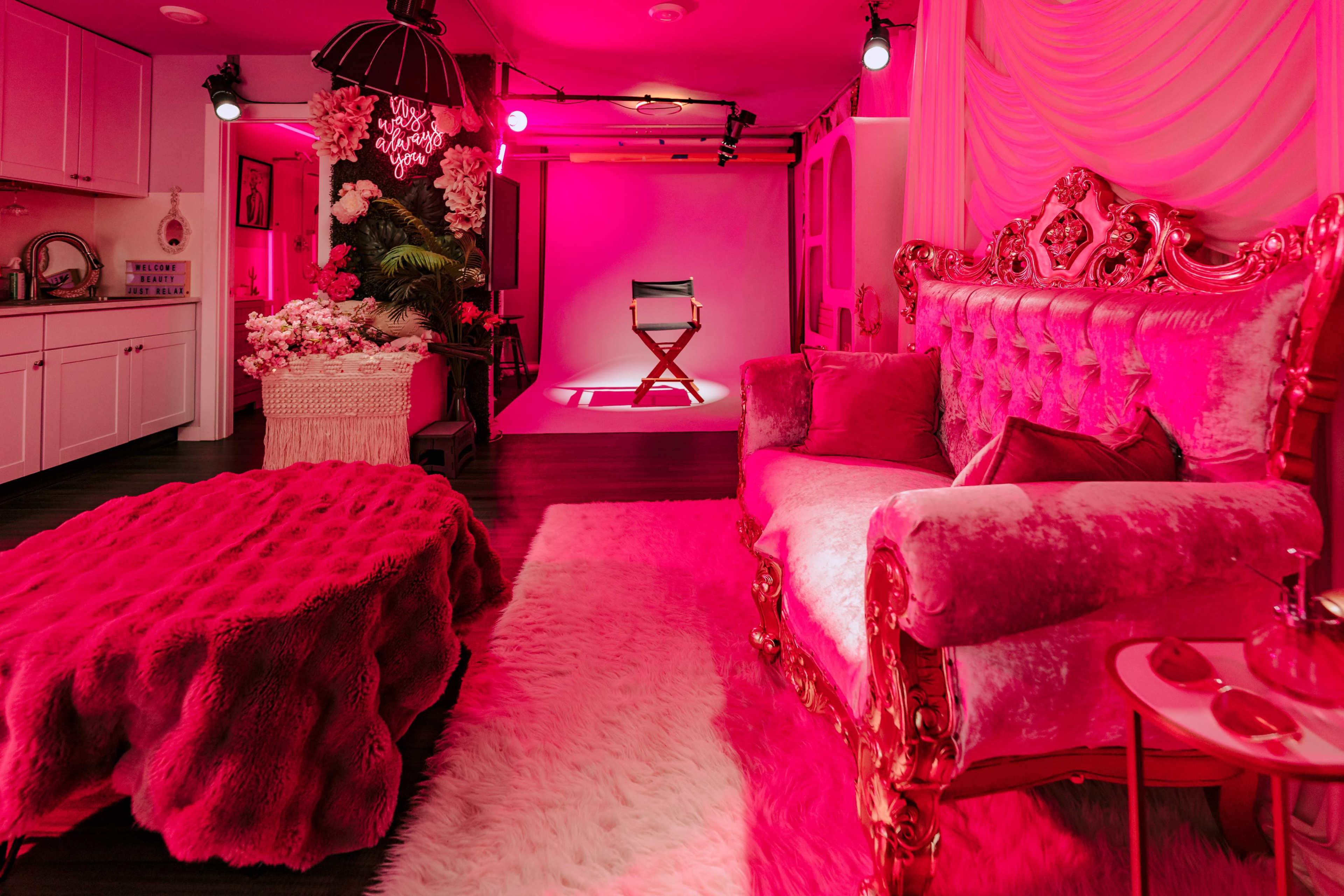 The image shows a stylish room featuring a plush pink couch, a decorative rug, and a spotlighted chair against a backdrop, all illuminated with pink lighting.