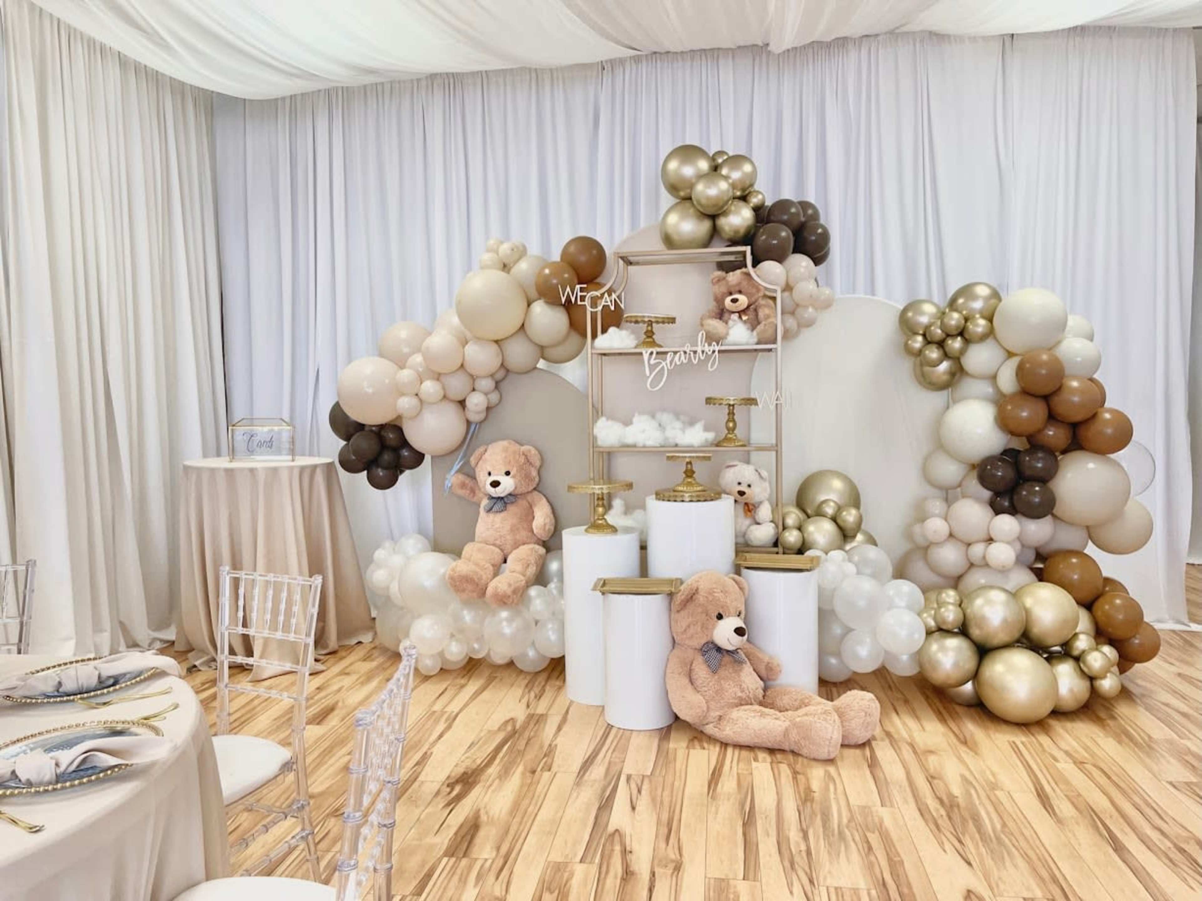 The image depicts a decorated event space featuring a backdrop of balloons in various shades, with plush teddy bears positioned in front of a tiered display.