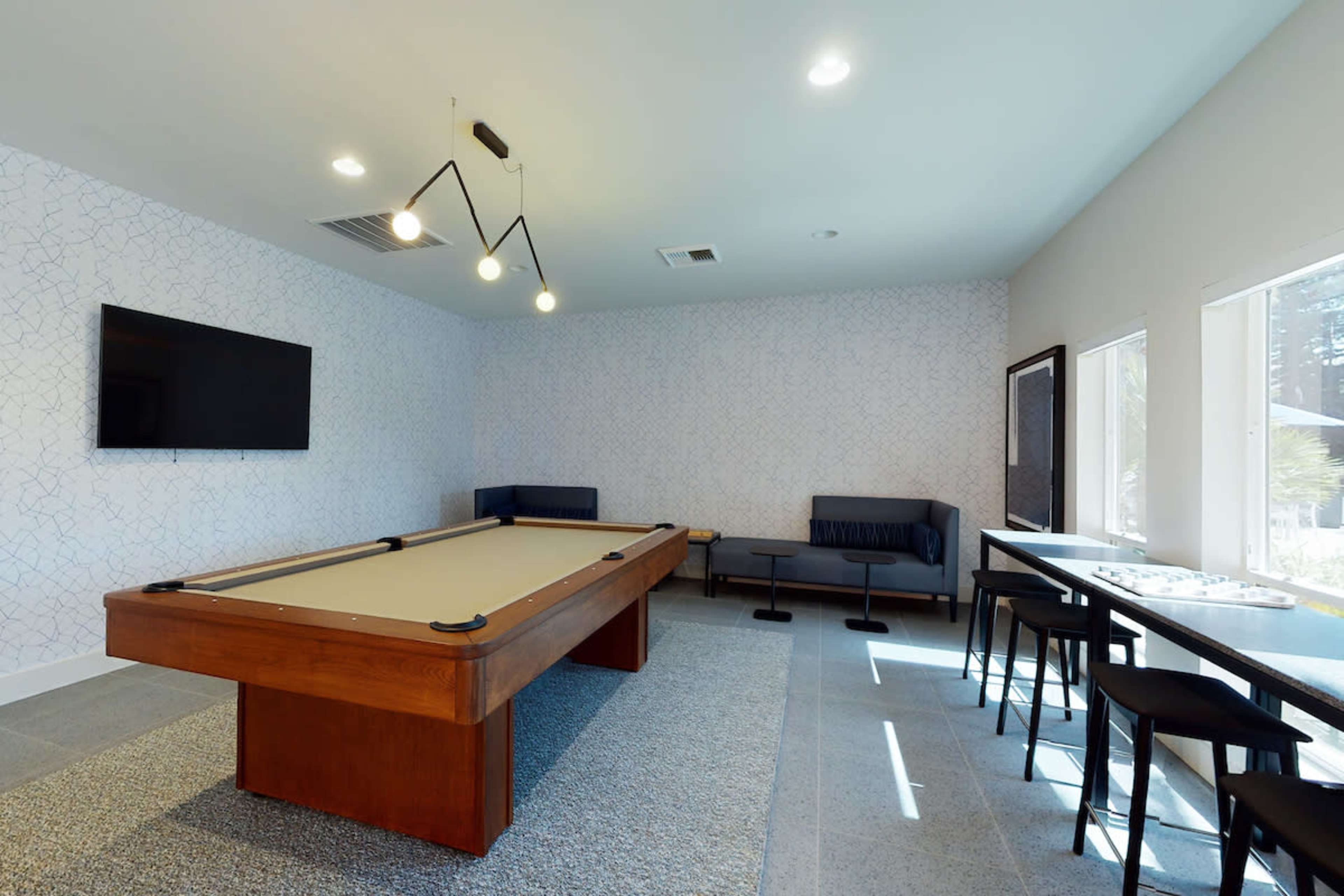 A room with a pool table, a wall-mounted television, and seating areas with modern furniture and large windows.