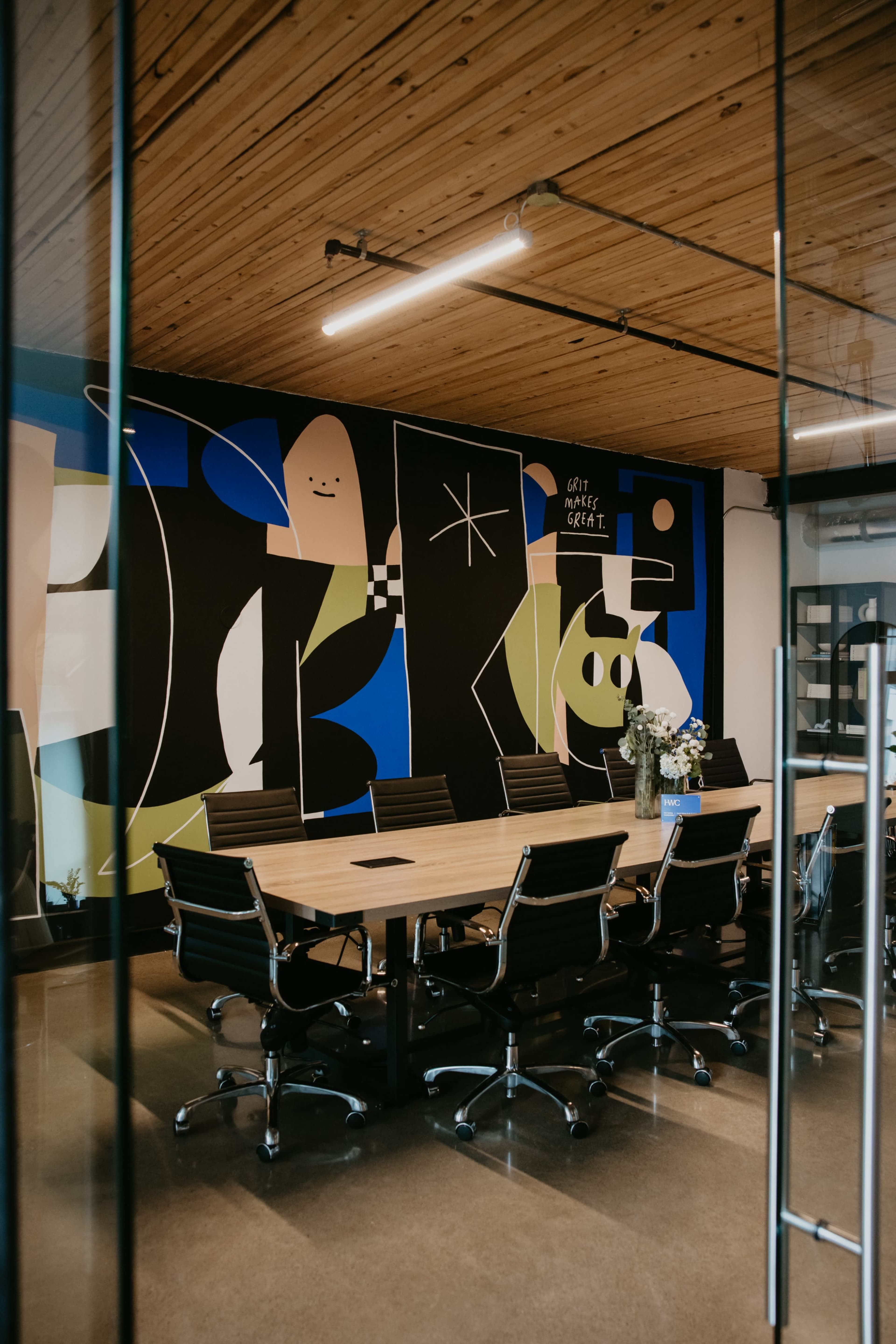 A modern conference room features a large wooden table surrounded by black swivel chairs, with a colorful abstract mural on the wall.