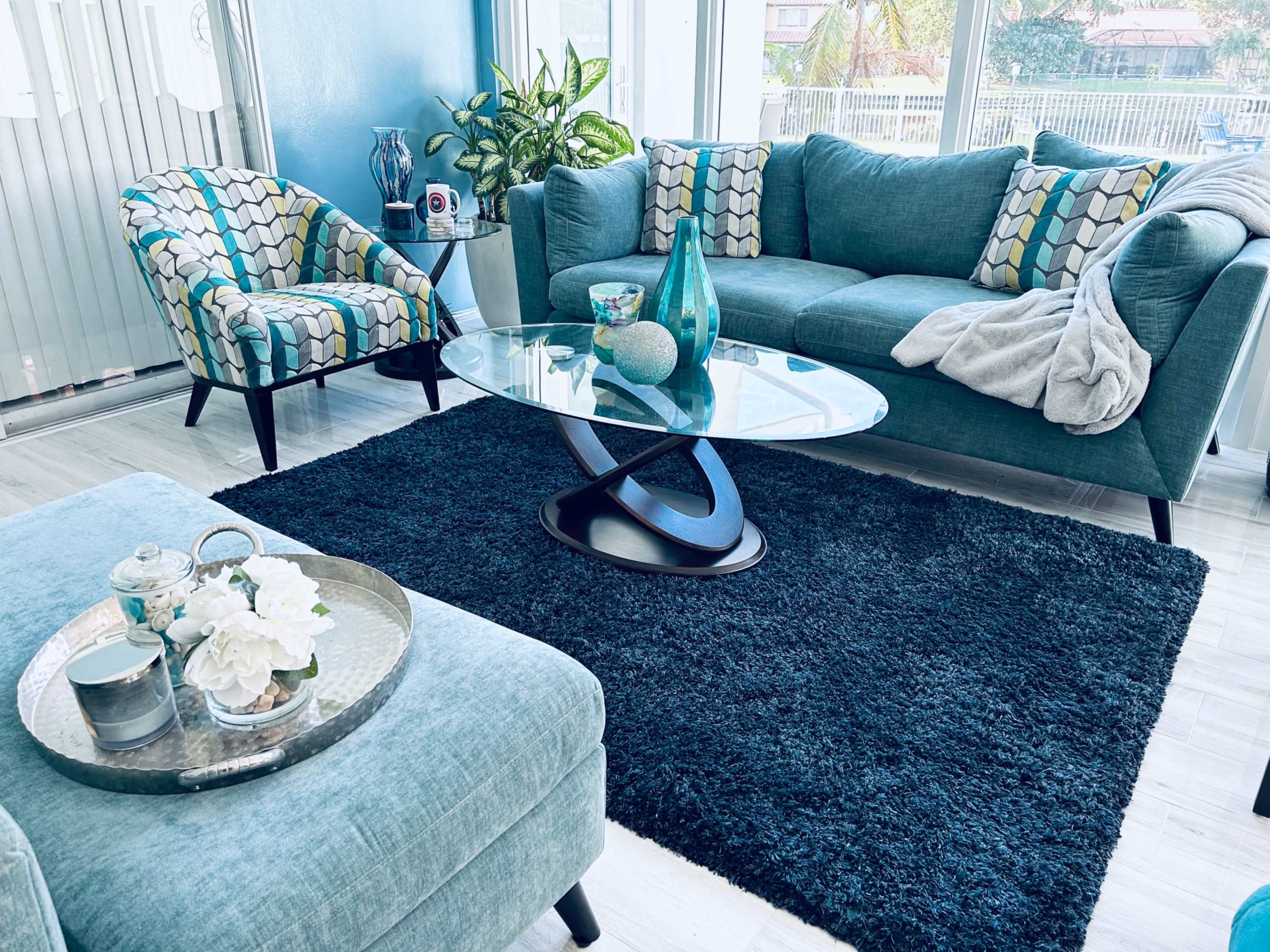 The living room features a teal sofa and chairs with patterned upholstery, a round glass coffee table, and a dark rug, surrounded by plants and natural light.