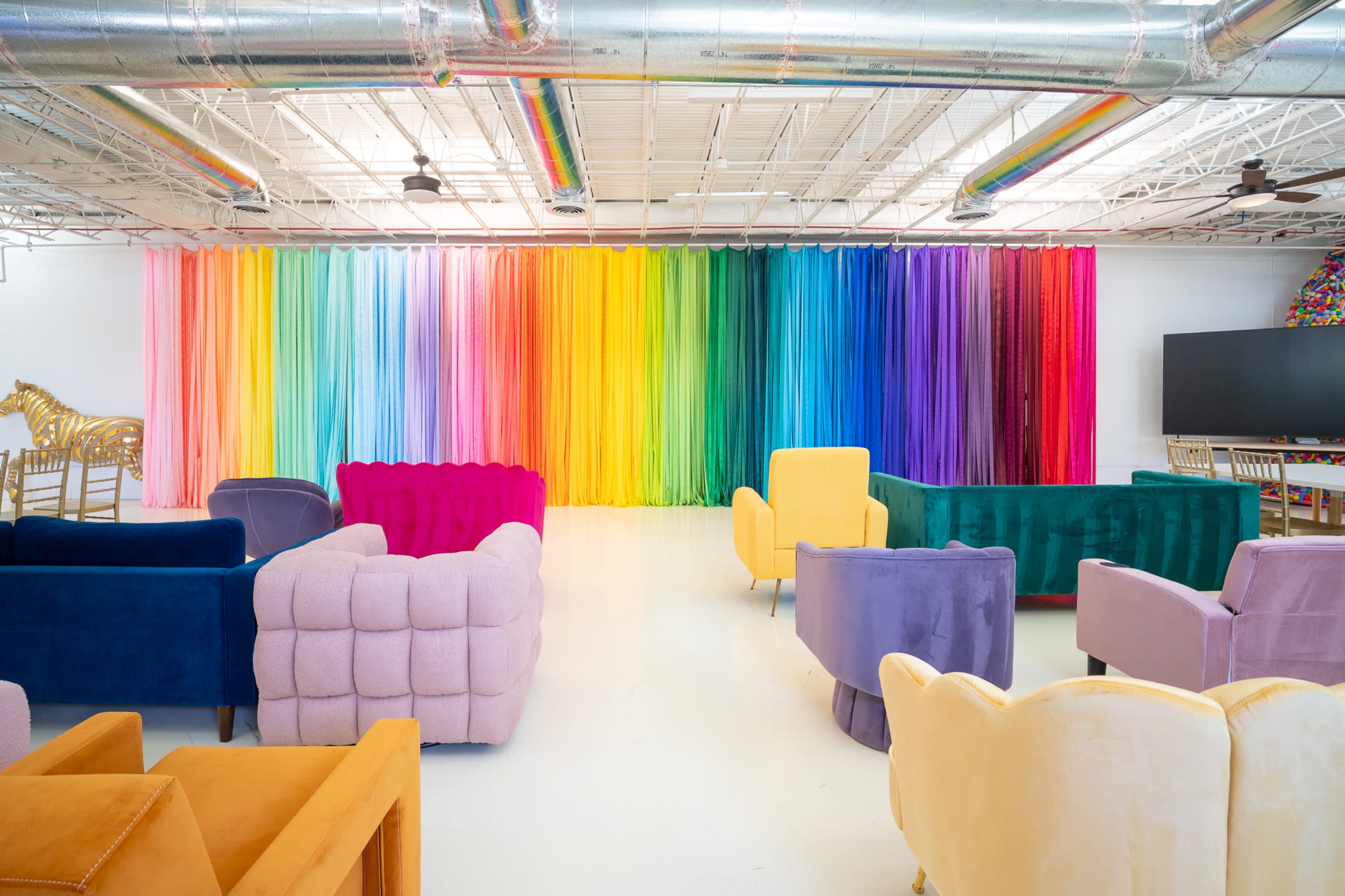 The image shows a spacious, colorful room with various velvet chairs arranged in multiple seating areas, against a backdrop of cascading rainbow fabric.
