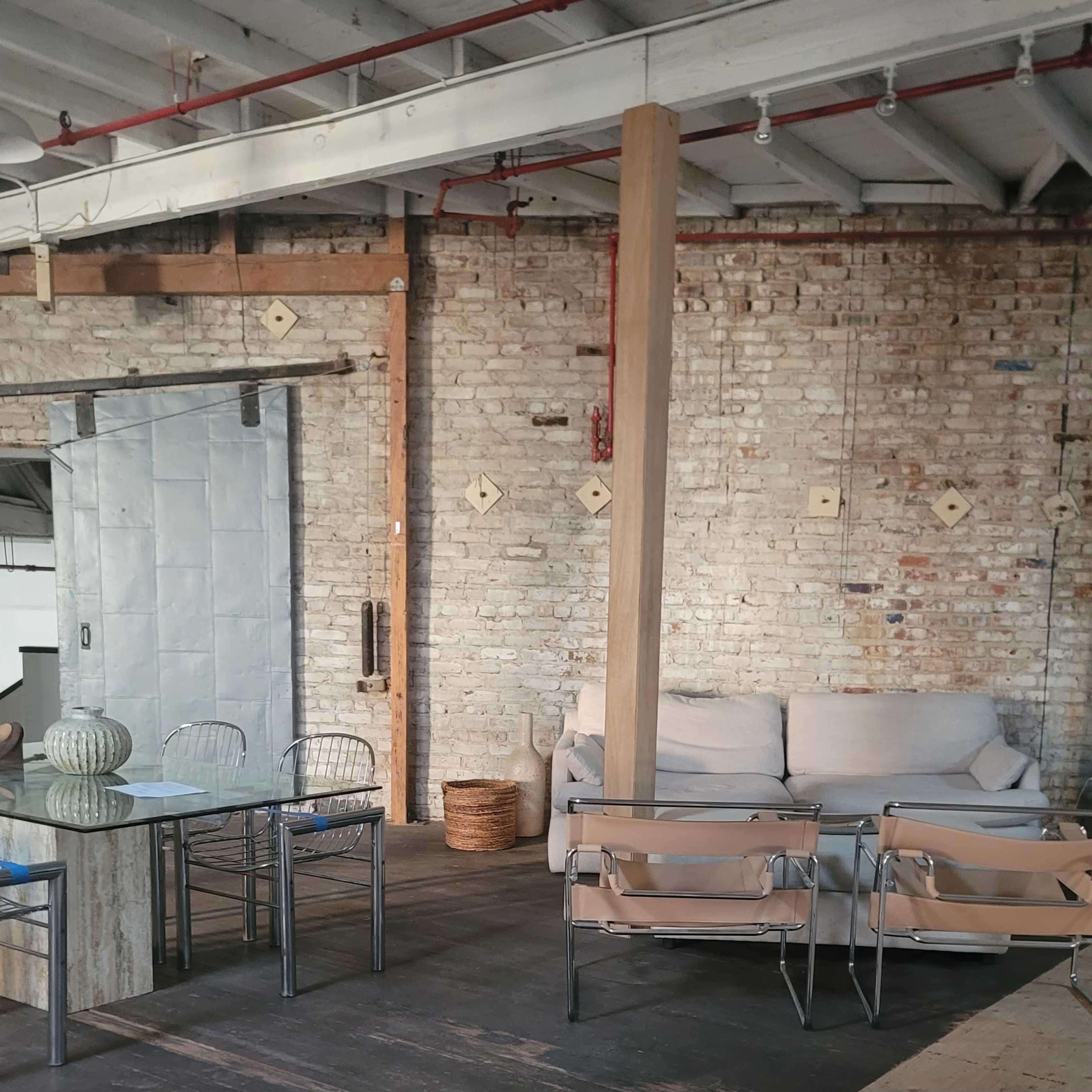 The image shows a spacious interior with exposed brick walls, a wooden beam, a dining table with metal chairs, a sofa, and a woven basket.