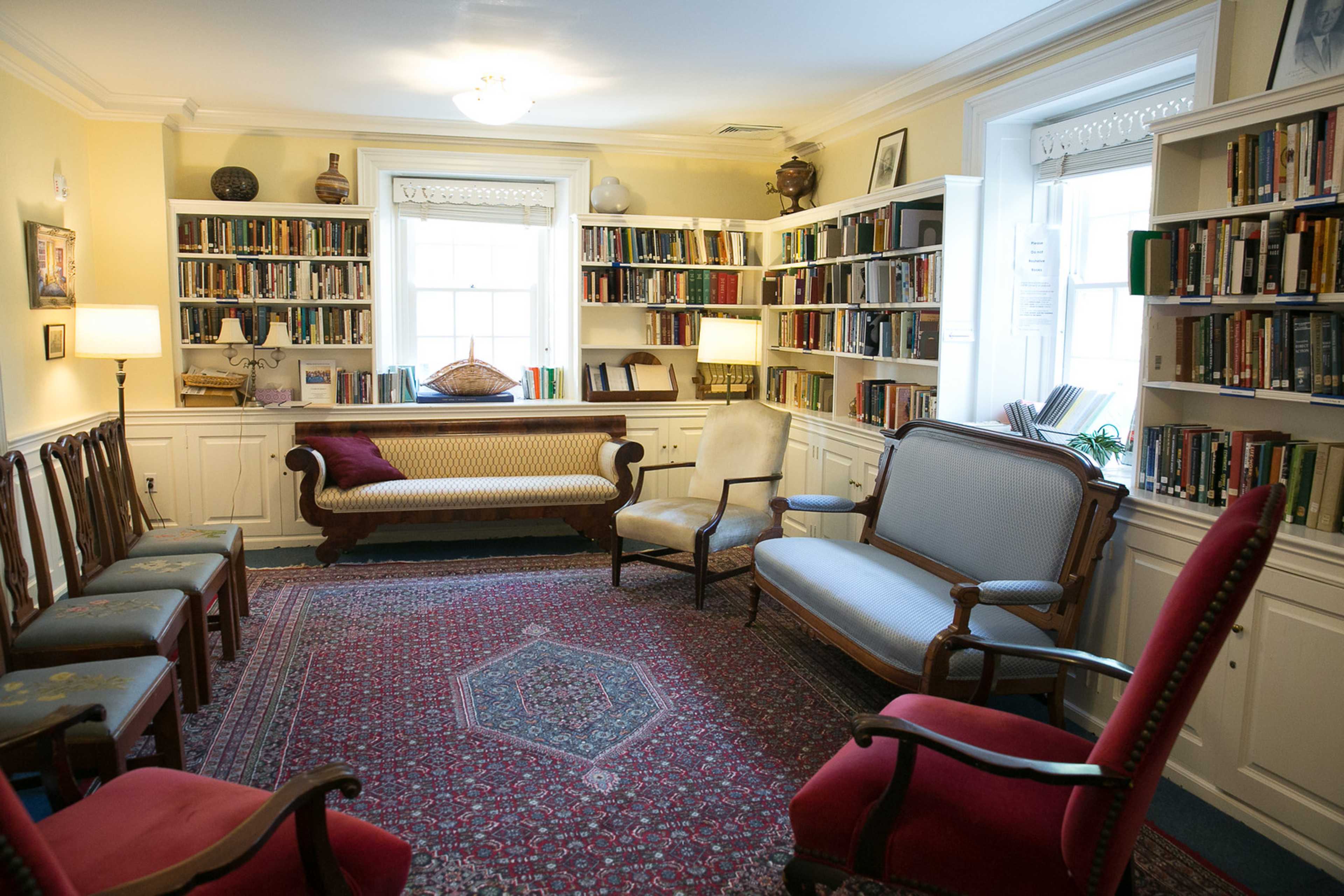 A cozy library with bookshelves lining the walls, a patterned rug on the floor, and several upholstered chairs arranged around a central seating area.