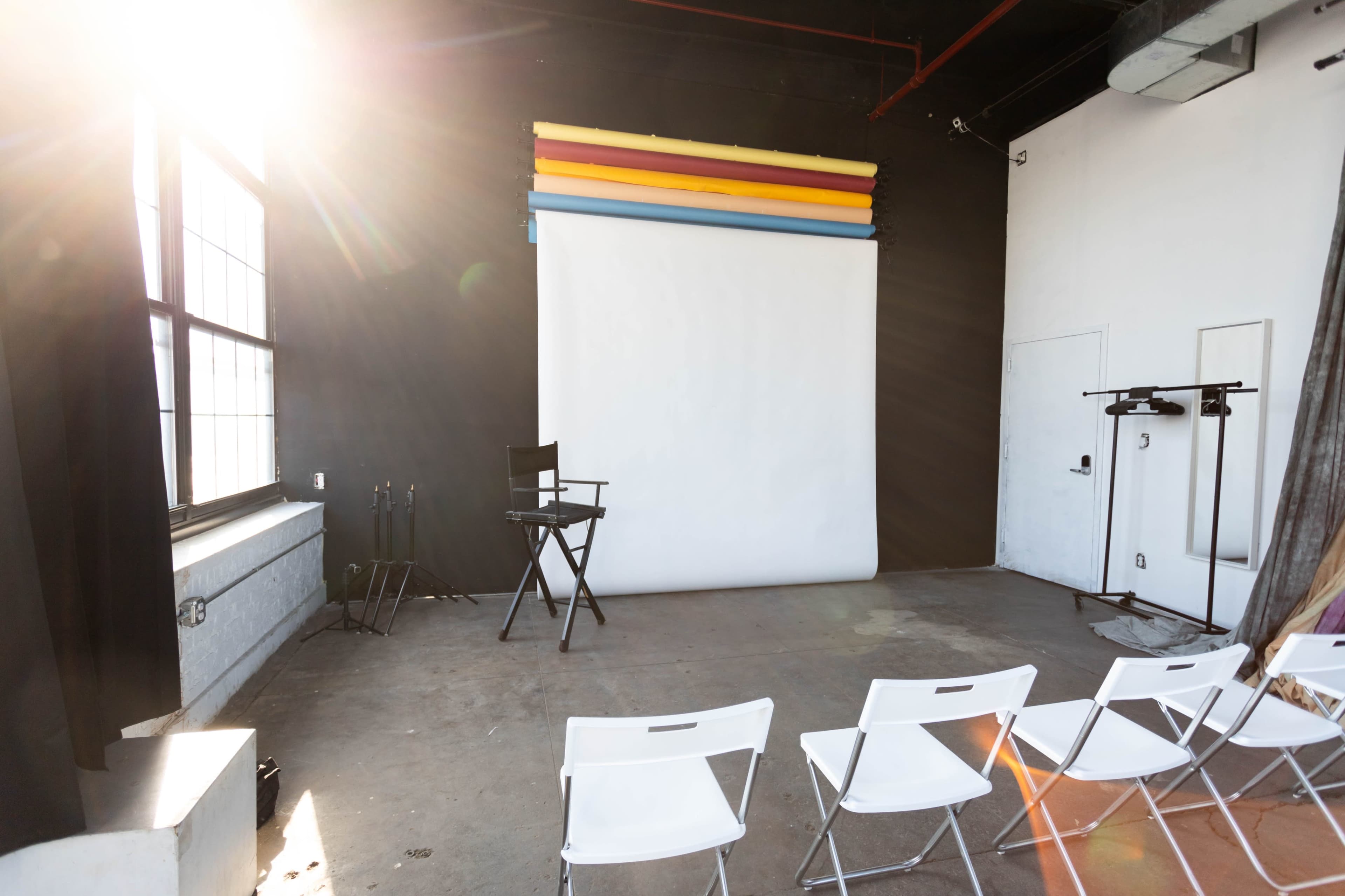 A photo studio with a white backdrop, colorful rolls of paper above it, a director's chair, and several white folding chairs arranged in front.
