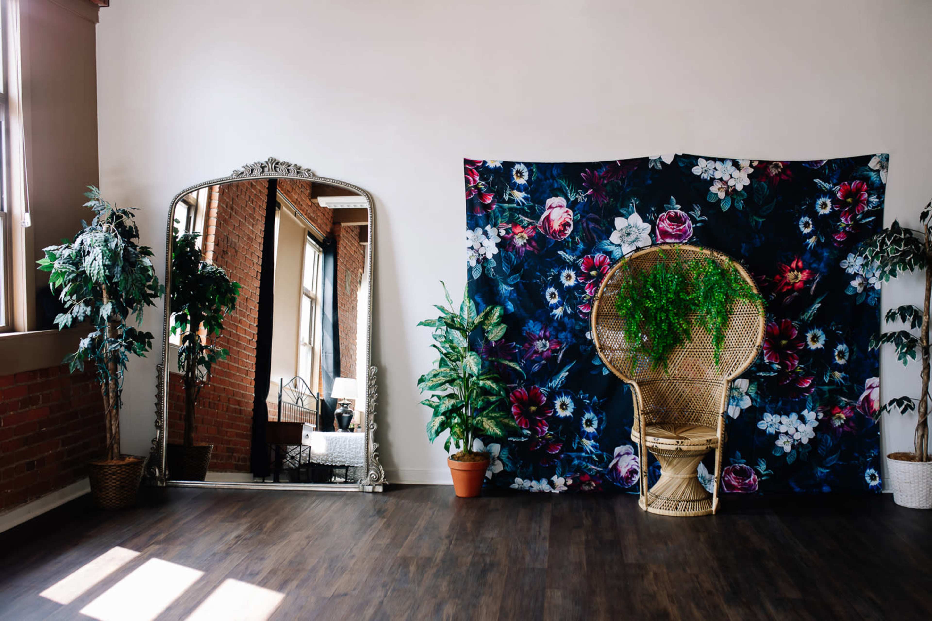 The room features a large decorative mirror, a floral backdrop, plants in pots, and a rattan chair.